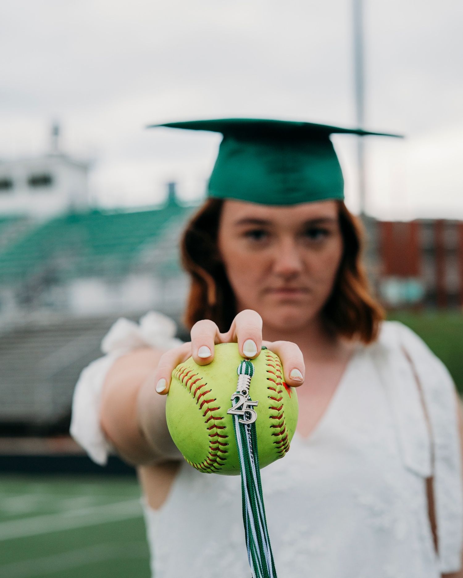 Female graduate in green cap holds a softball with a graduation tassel at a stadium.