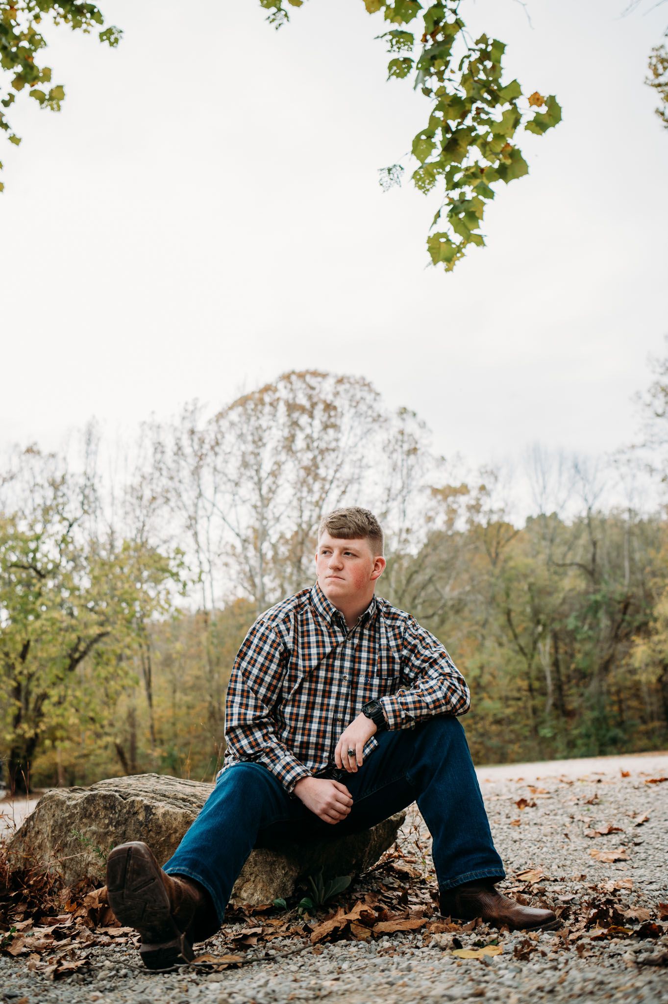 Young man in plaid shirt and jeans sitting on a rock surrounded by autumn leaves in a wooded outdoor setting.