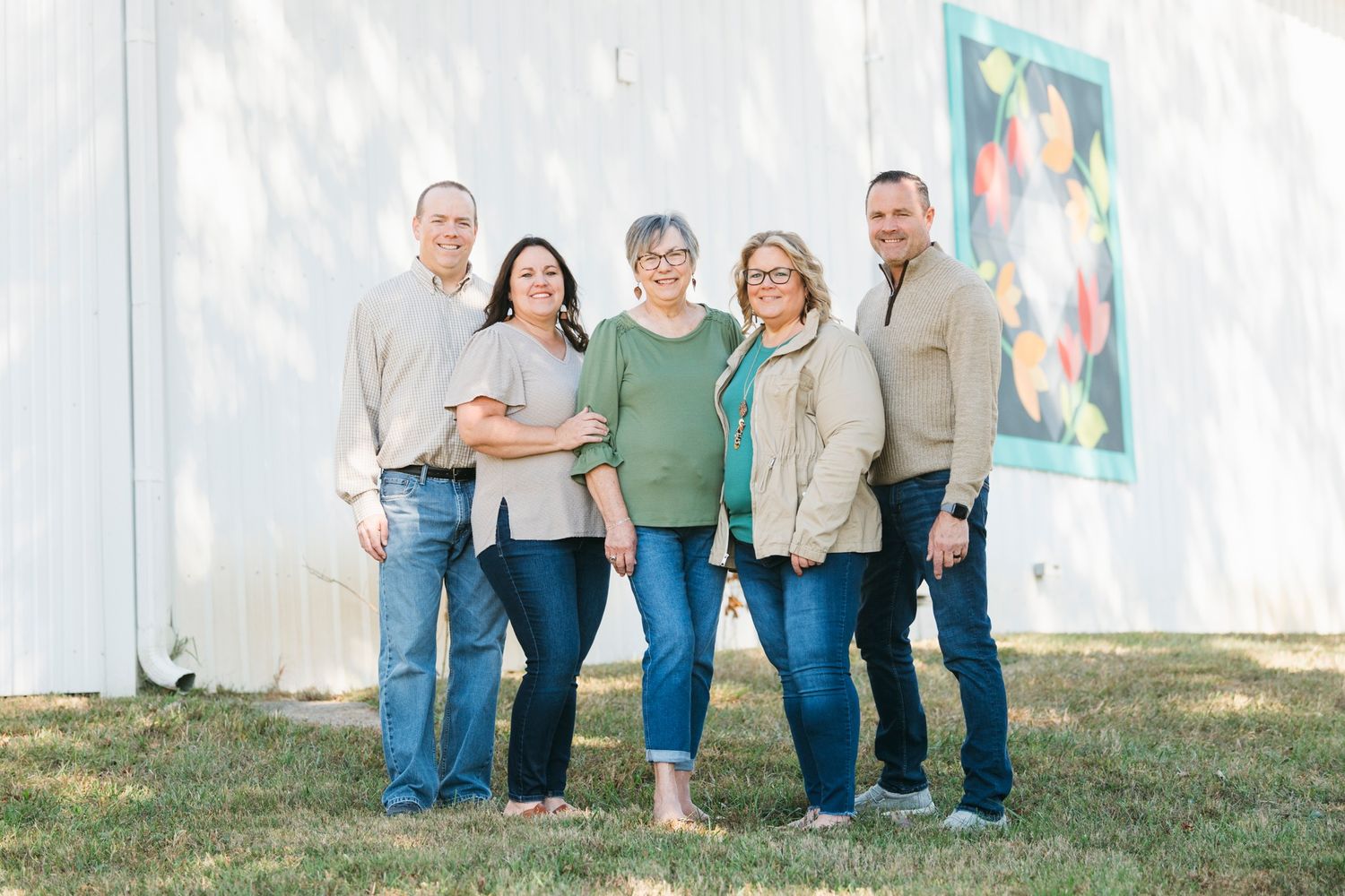 Group of five adults smiling together outdoors in front of white barn wall with colorful quilt.