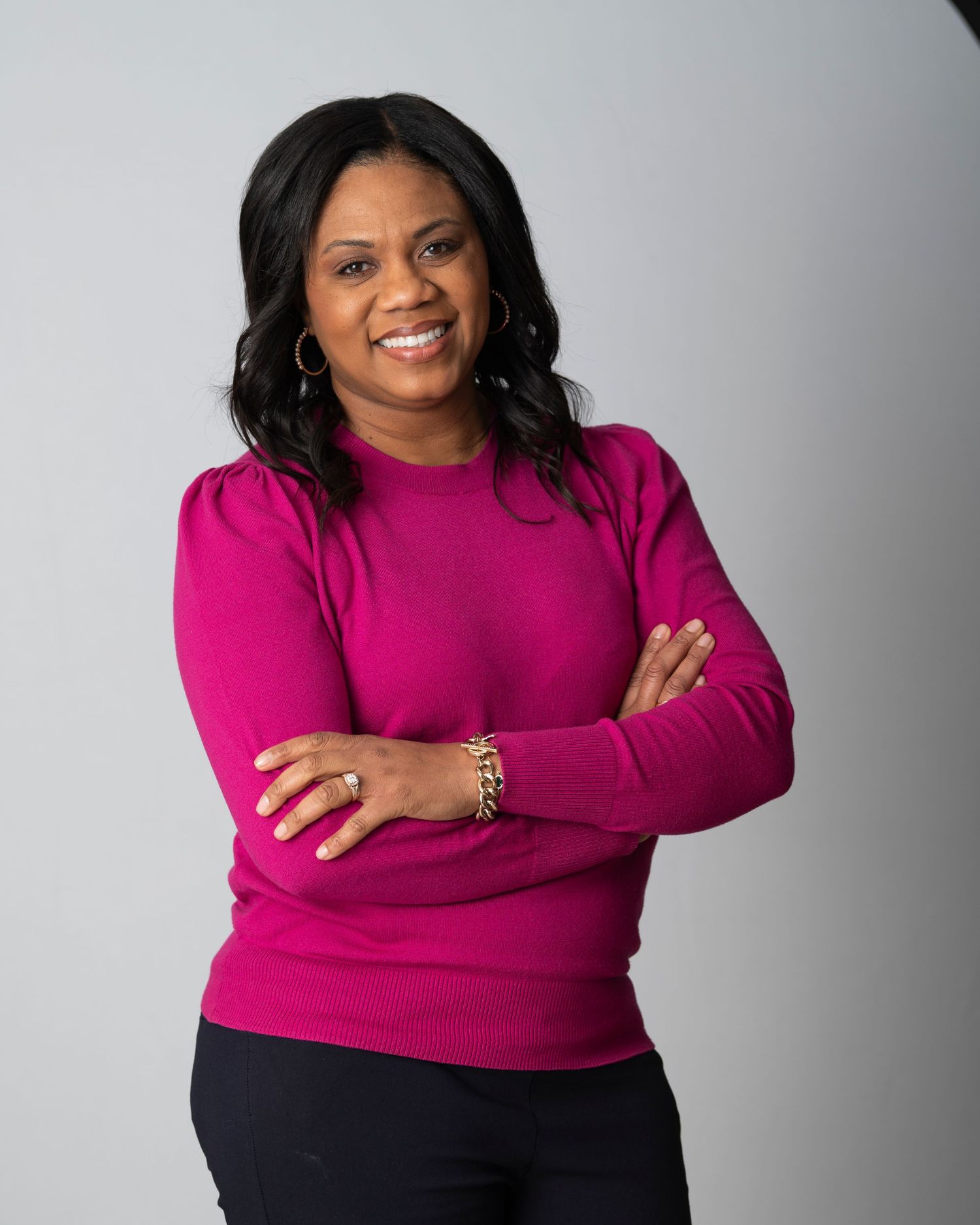 A confident woman in a magenta sweater poses with arms crossed and a bright smile against a gray studio background.