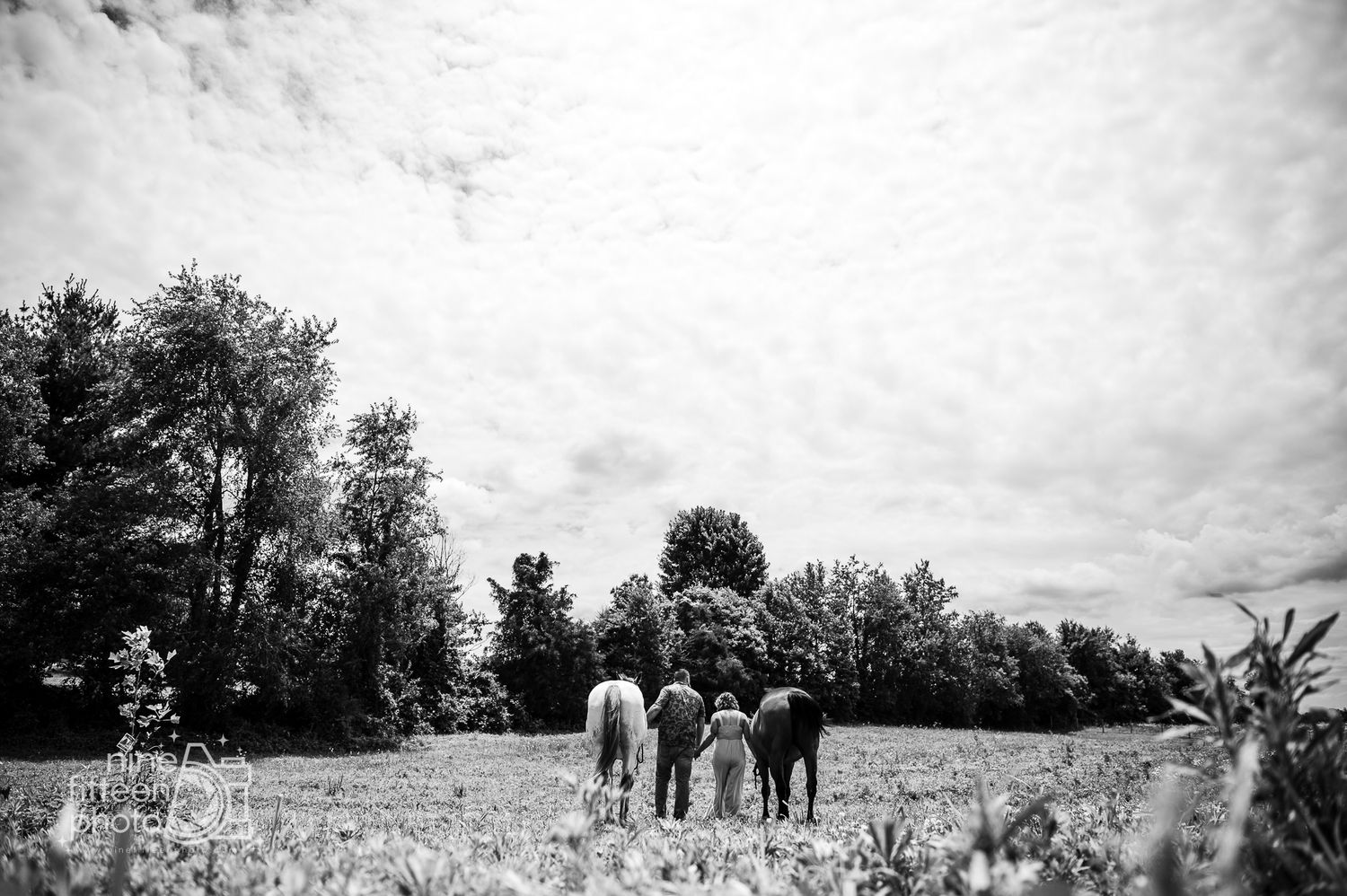 Black and white photo of two people standing with horses in a lush green field surrounded by trees.