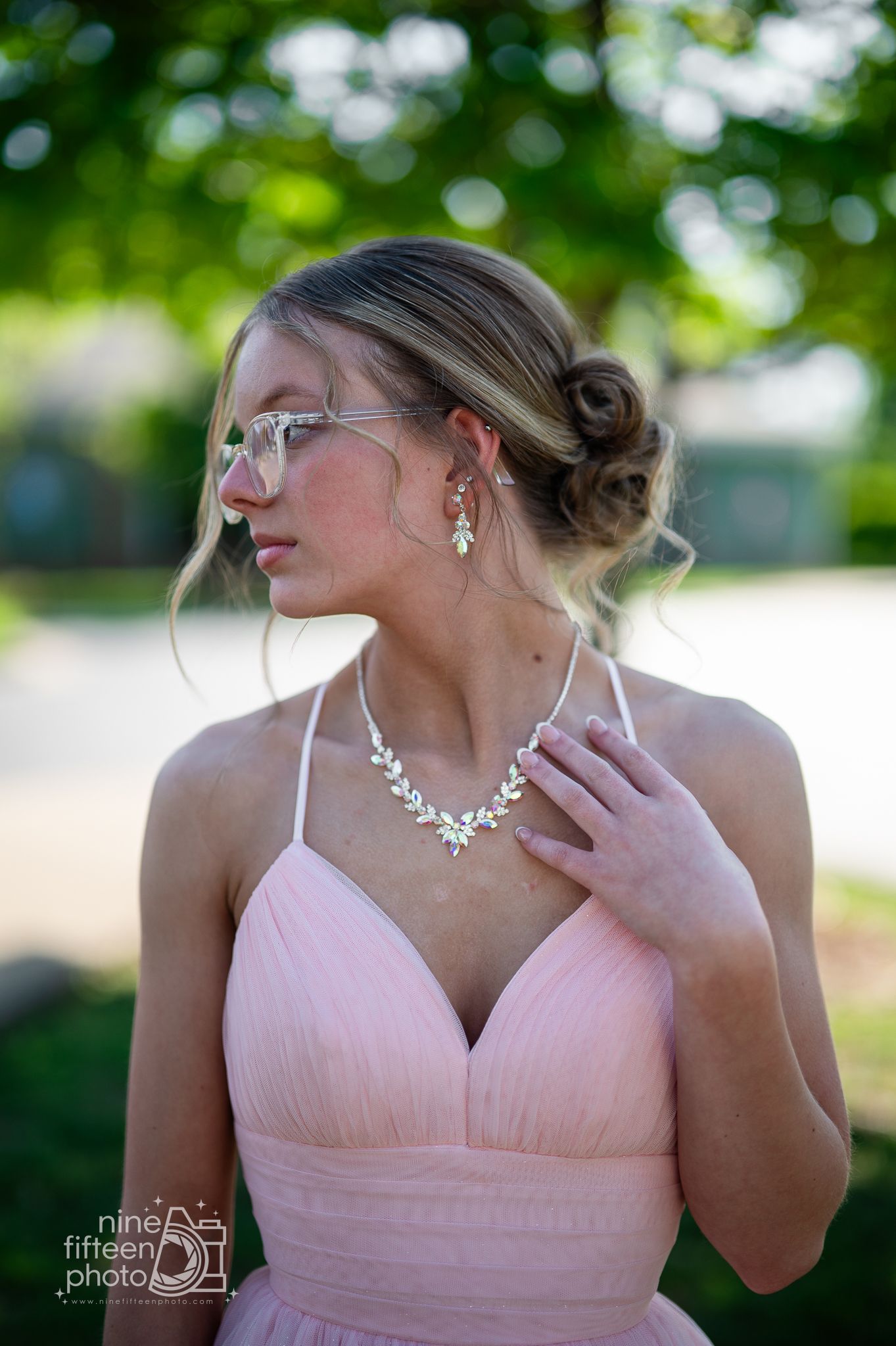 Young woman in pink gown with crystal necklace and earrings, wearing mirrored sunglasses at an outdoor event.