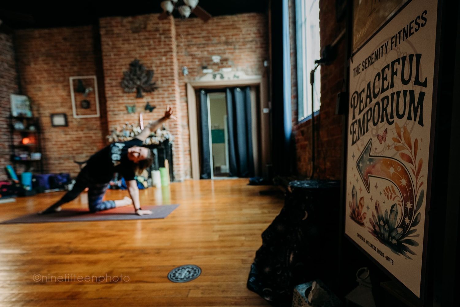 Yogi performs side angle pose on wood floor in brick-walled studio beside Peaceful Emporium sign.