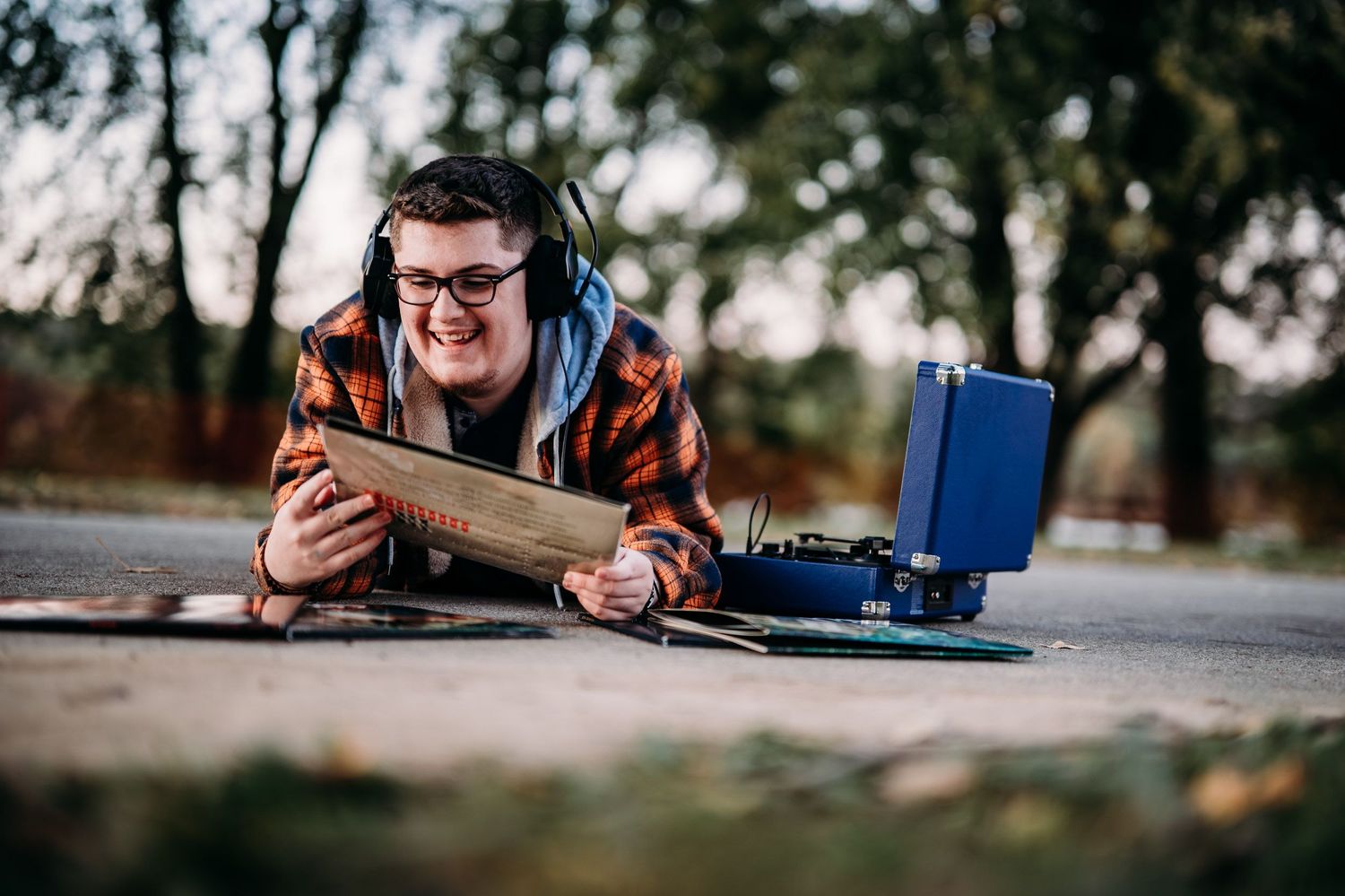 Young man with headphones lying on pavement smiling while holding a vinyl record next to a blue record player.