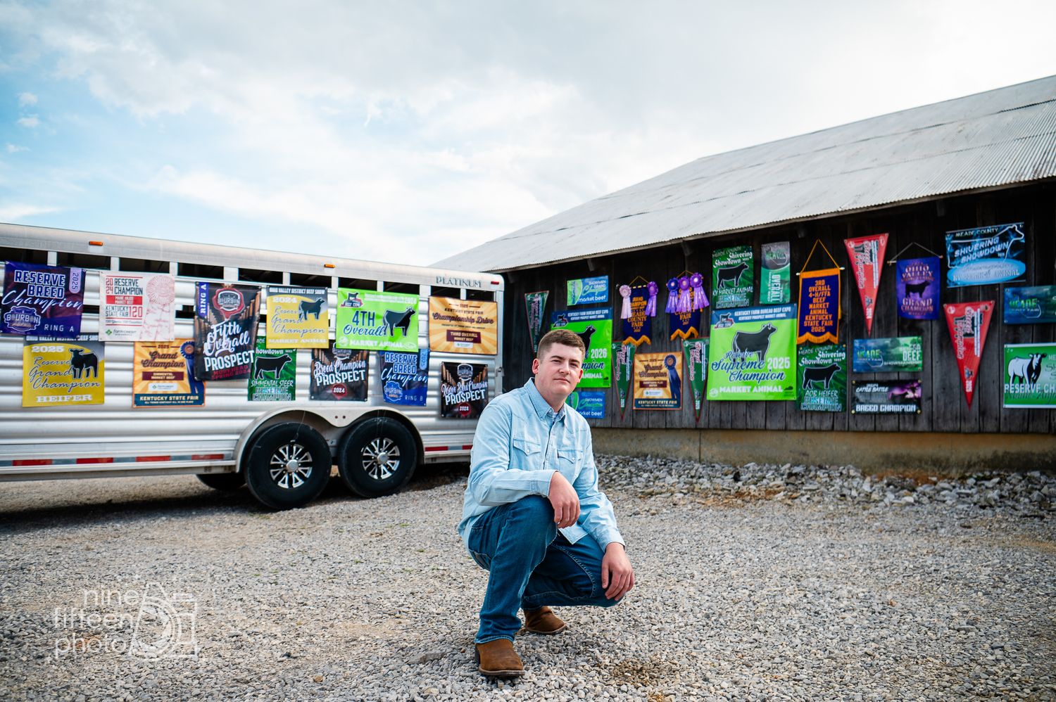 Young man crouching in front of a colorful poster-covered barn wall and trailer in an outdoor setting.