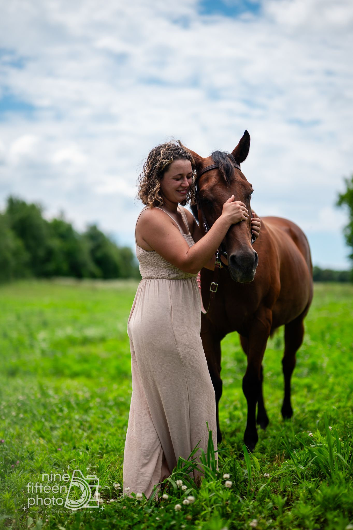 A woman in a beige dress affectionately embraces a brown horse in a lush green field under a cloudy sky.