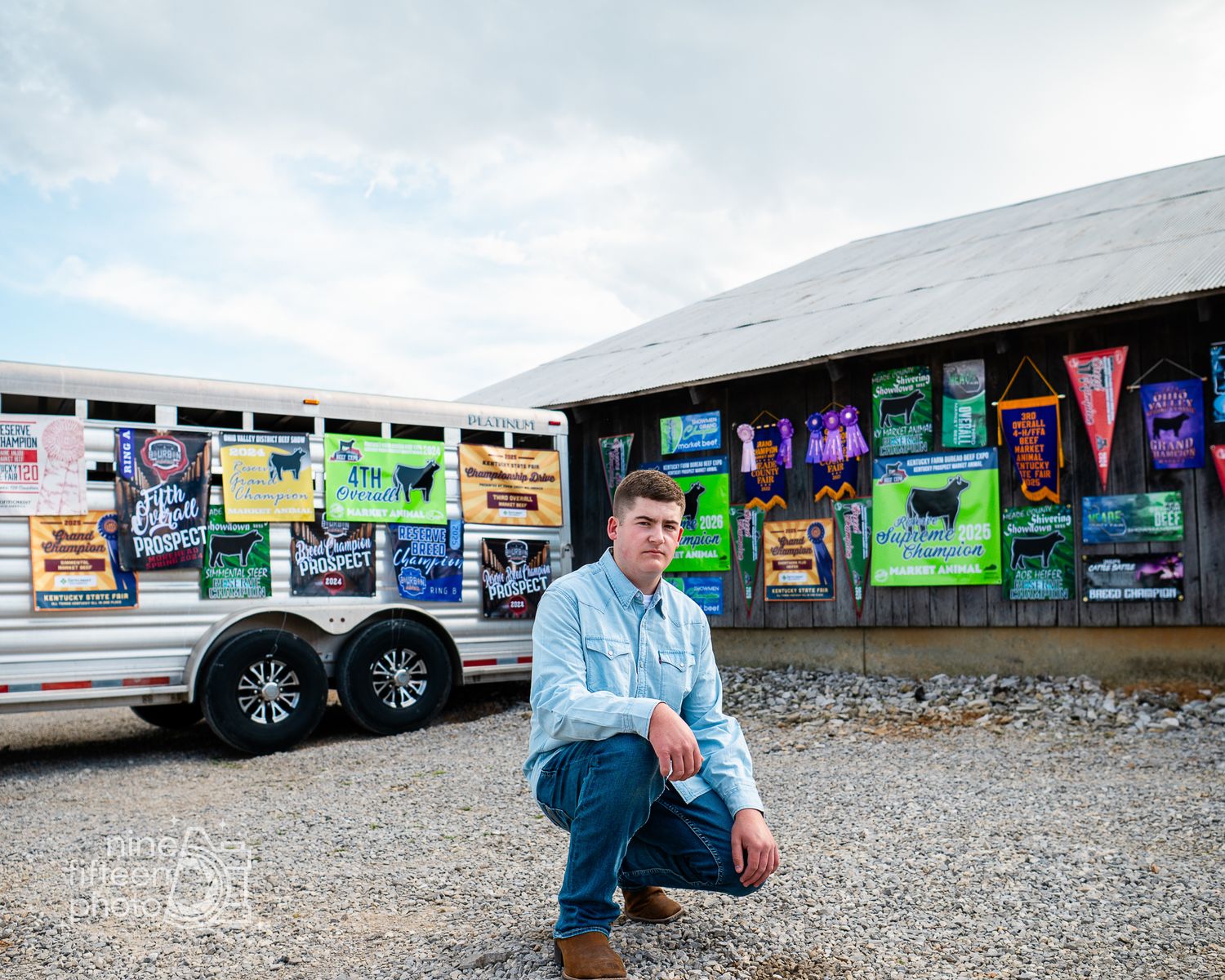 Young man in denim shirt crouching near colorful barn and trailer with event posters at an outdoor venue.