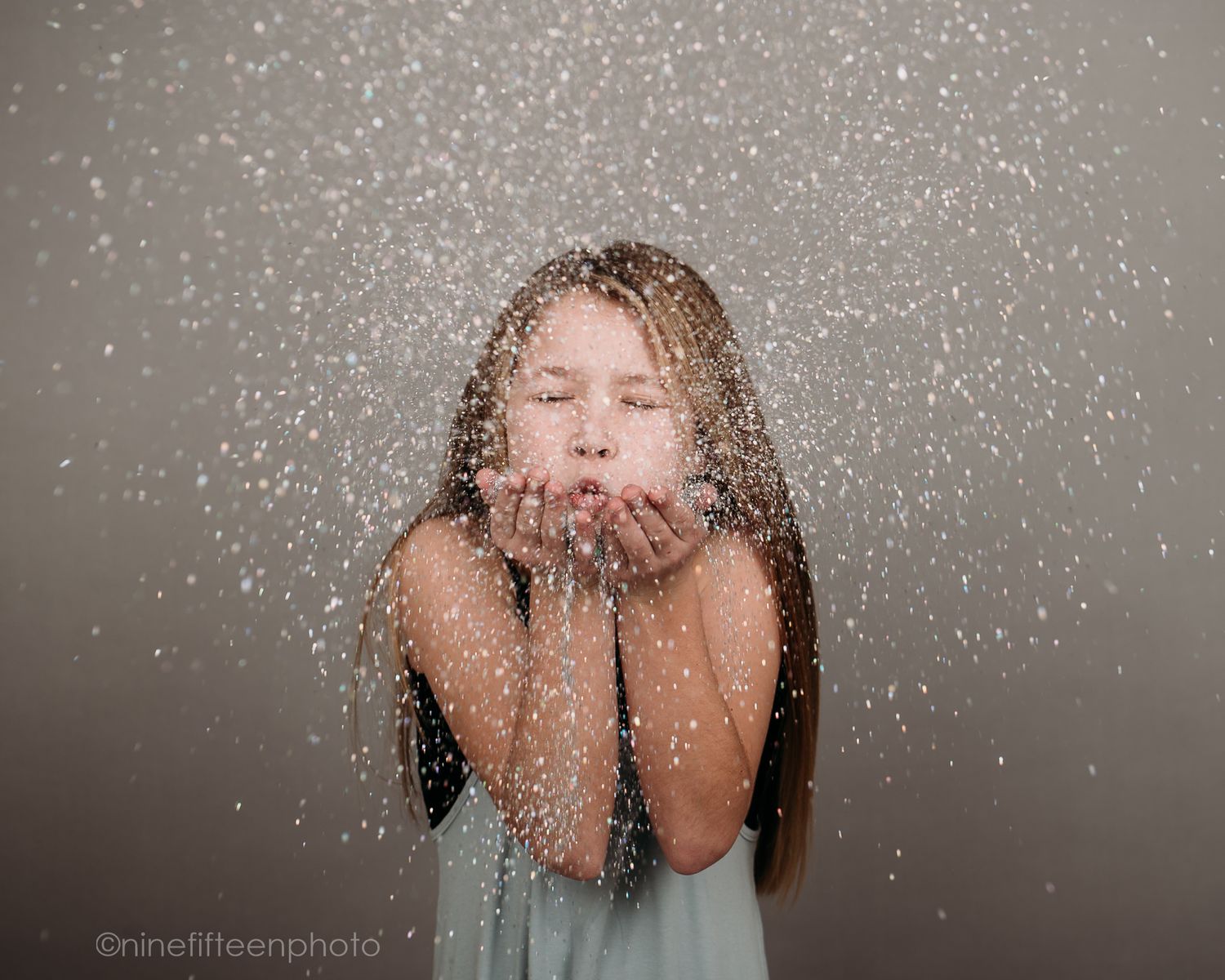 Young girl blowing sparkling glitter from cupped hands against a neutral gray background.