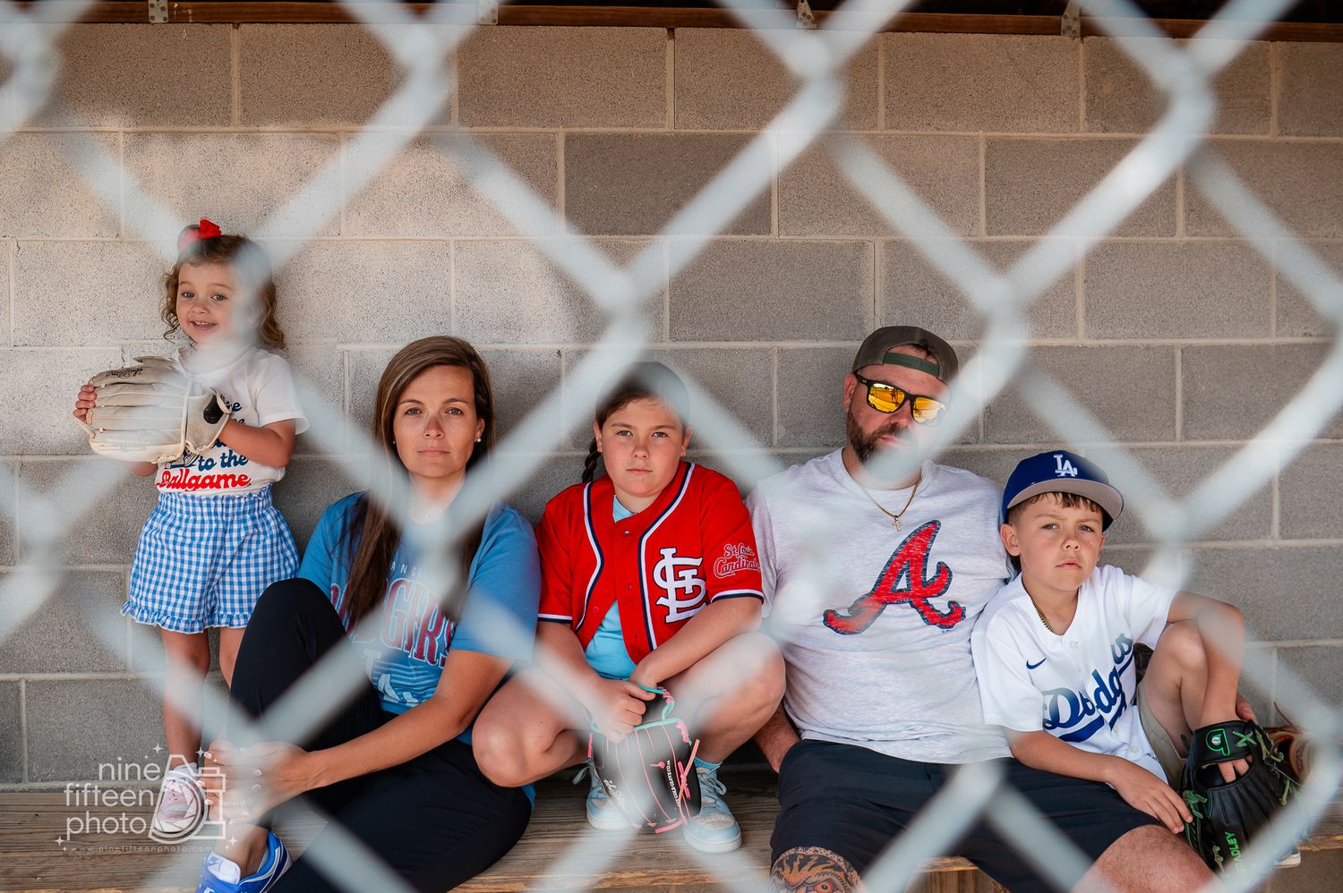 Family of five sitting against a wall, viewed through chain-link fence, wearing baseball jerseys at a sports event.