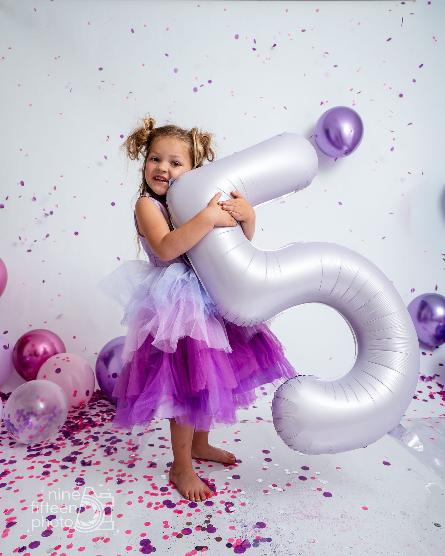 A smiling girl in a purple tutu hugs a large number 5 balloon surrounded by confetti and balloons for her 5th birthday.