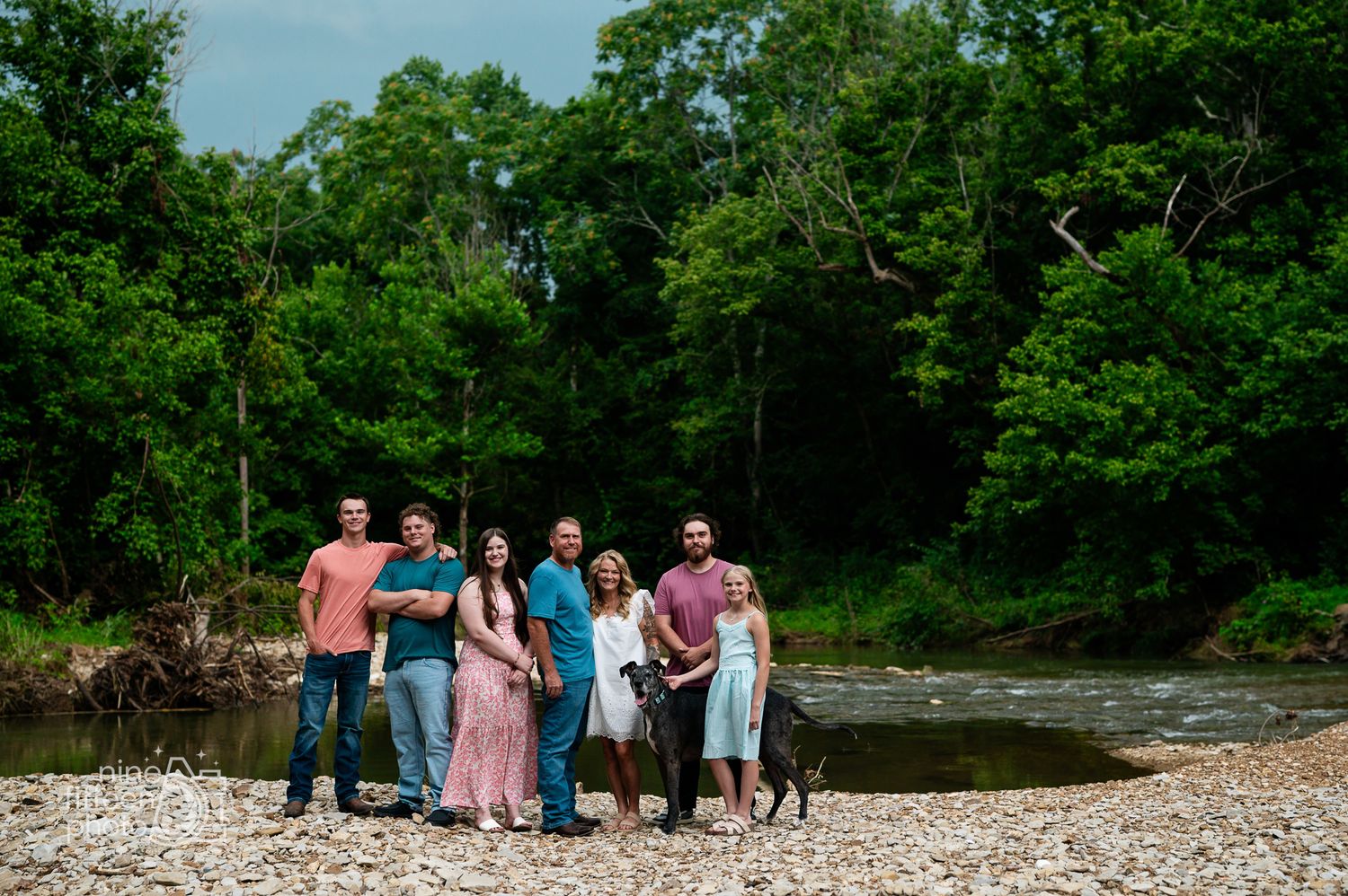 Family group portrait on rocky riverbank with lush green forest backdrop on a cloudy day.