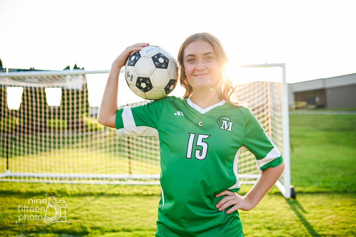 Female soccer player in green jersey number 15 holds ball on shoulder in front of goal net on sunny field.