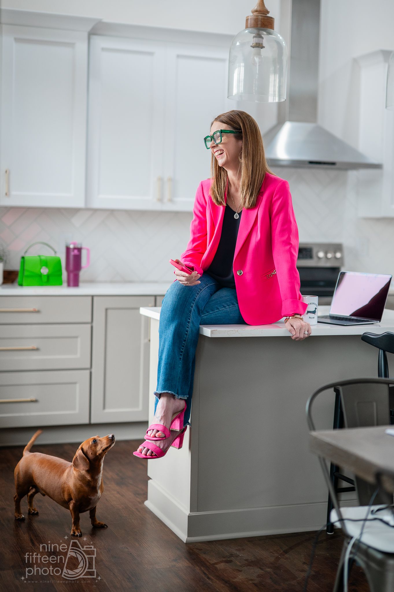 Woman in pink blazer and heels sits on kitchen counter smiling, with a dachshund dog nearby and laptop on counter.