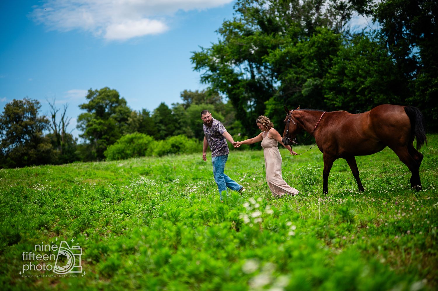 Couple holding hands walking through a lush green field beside a brown horse on a sunny day.