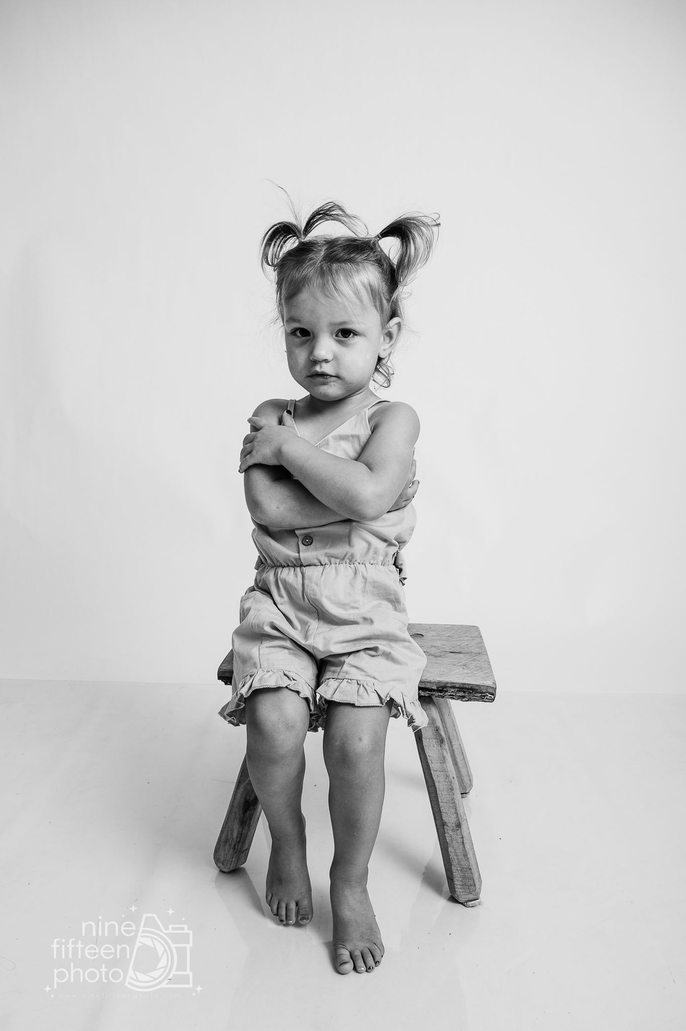 Black and white portrait of a toddler girl with pigtails sitting on a wooden stool, arms crossed, wearing a romper.