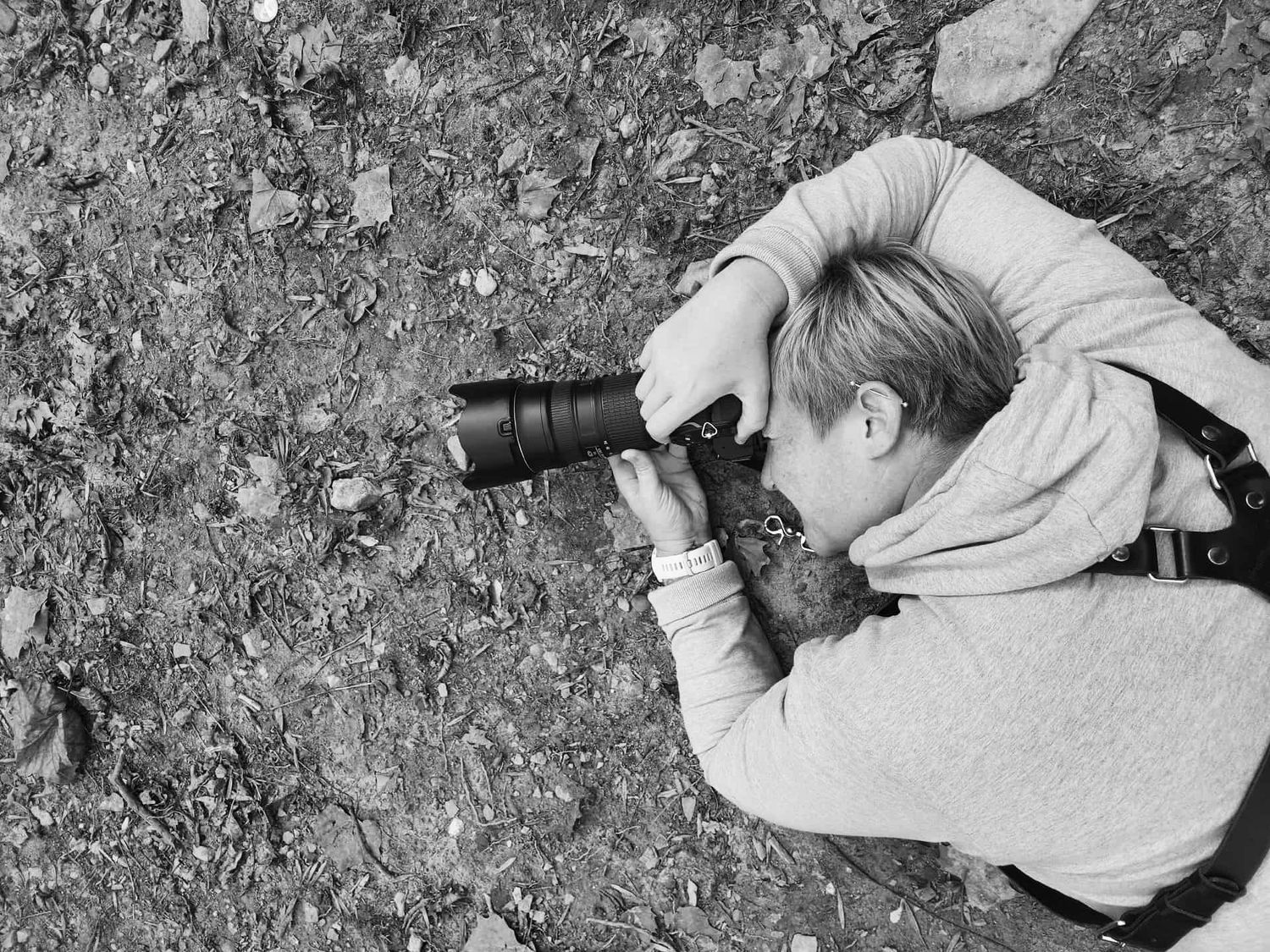 A black and white overhead shot of a photographer lying on the ground capturing a close-up nature photo.