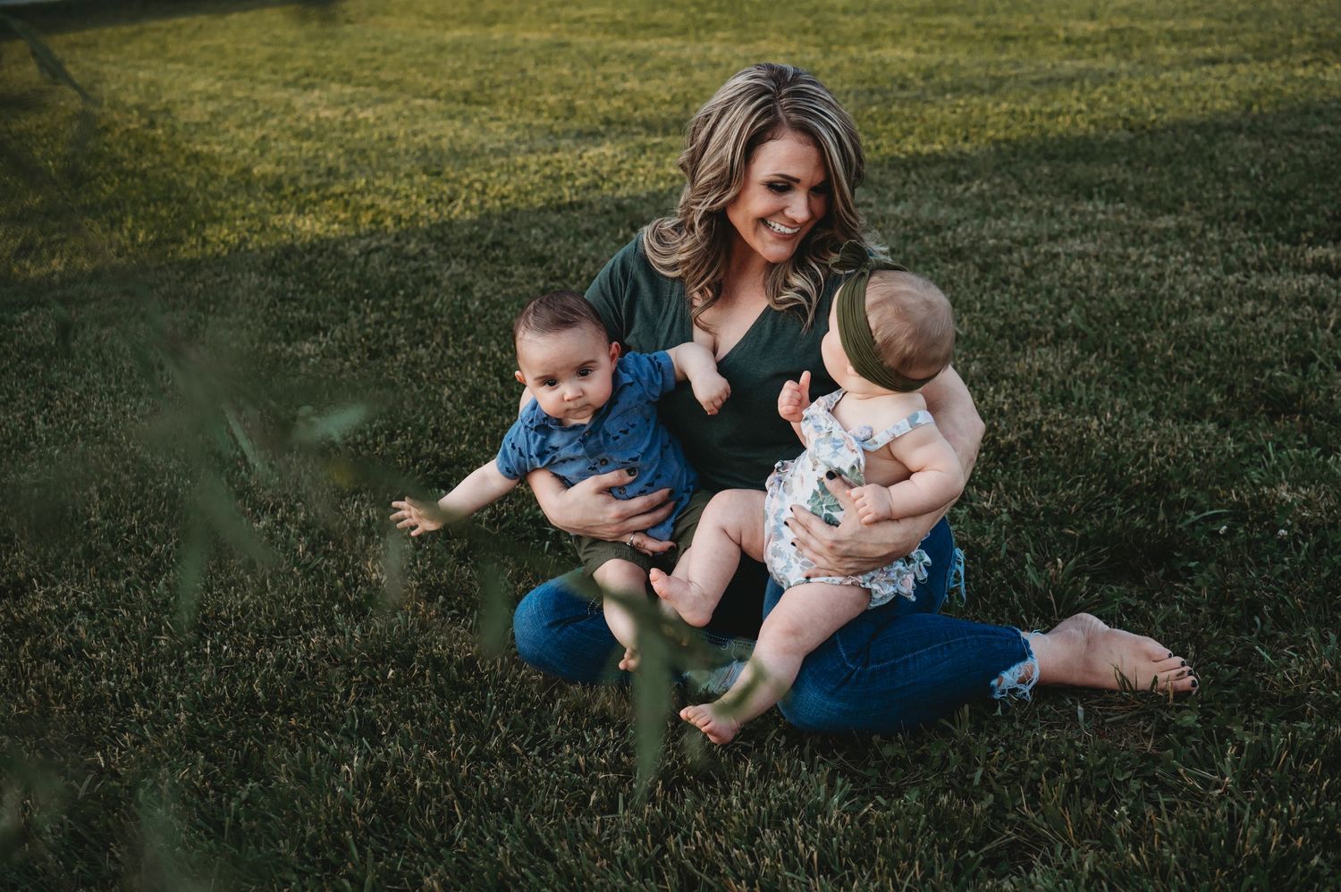Mother sitting on grass with two young children in casual outdoor family portrait.