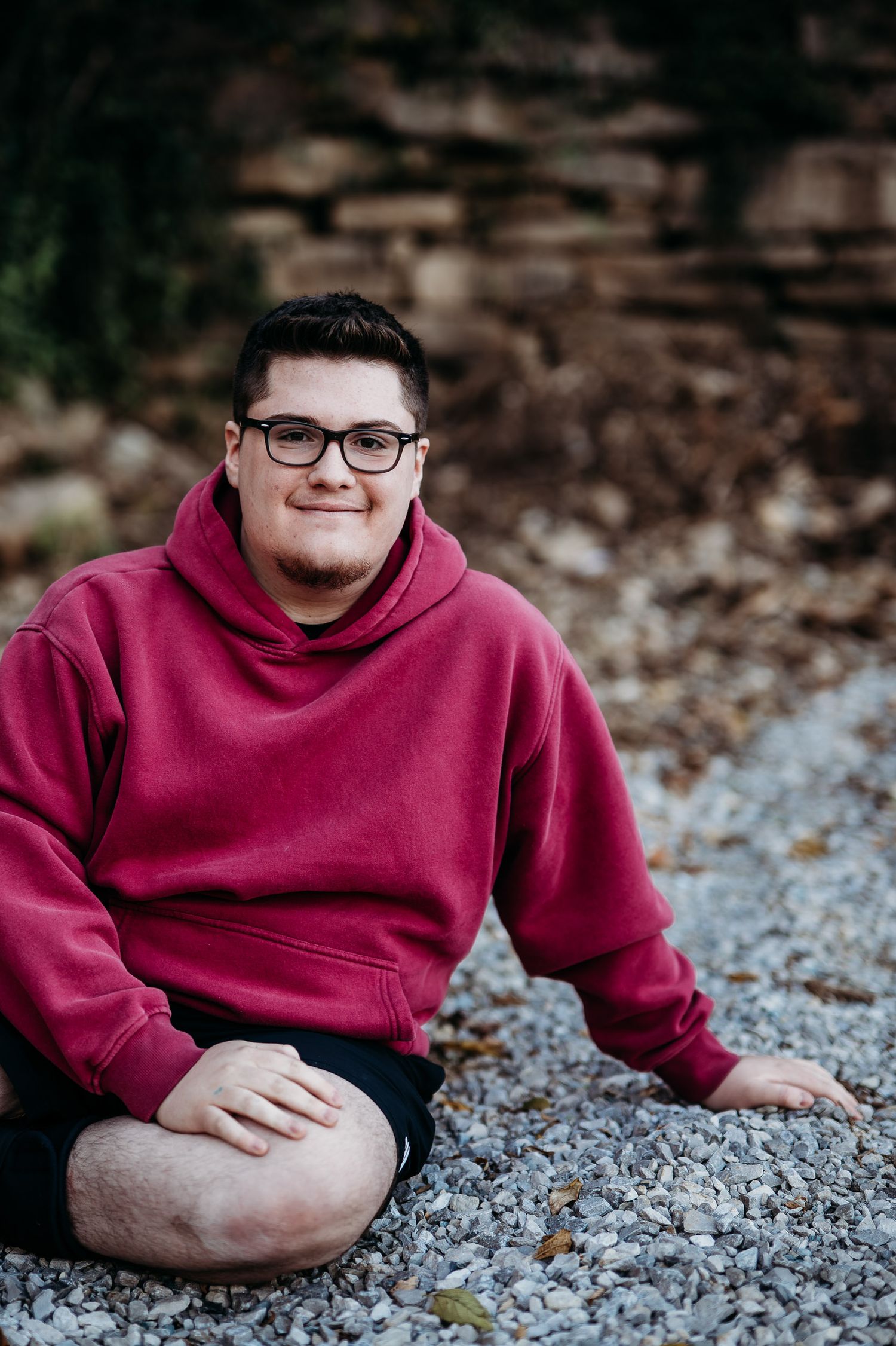 Luca sitting on gravel holding a football in his red hoodie during his MCHS senior session in Brandenburg KY.
