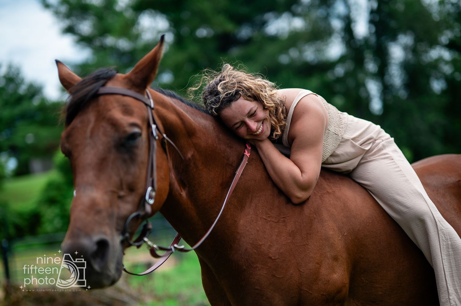 Woman in a beige dress lying on a chestnut horse, embracing it warmly in a lush green outdoor setting.