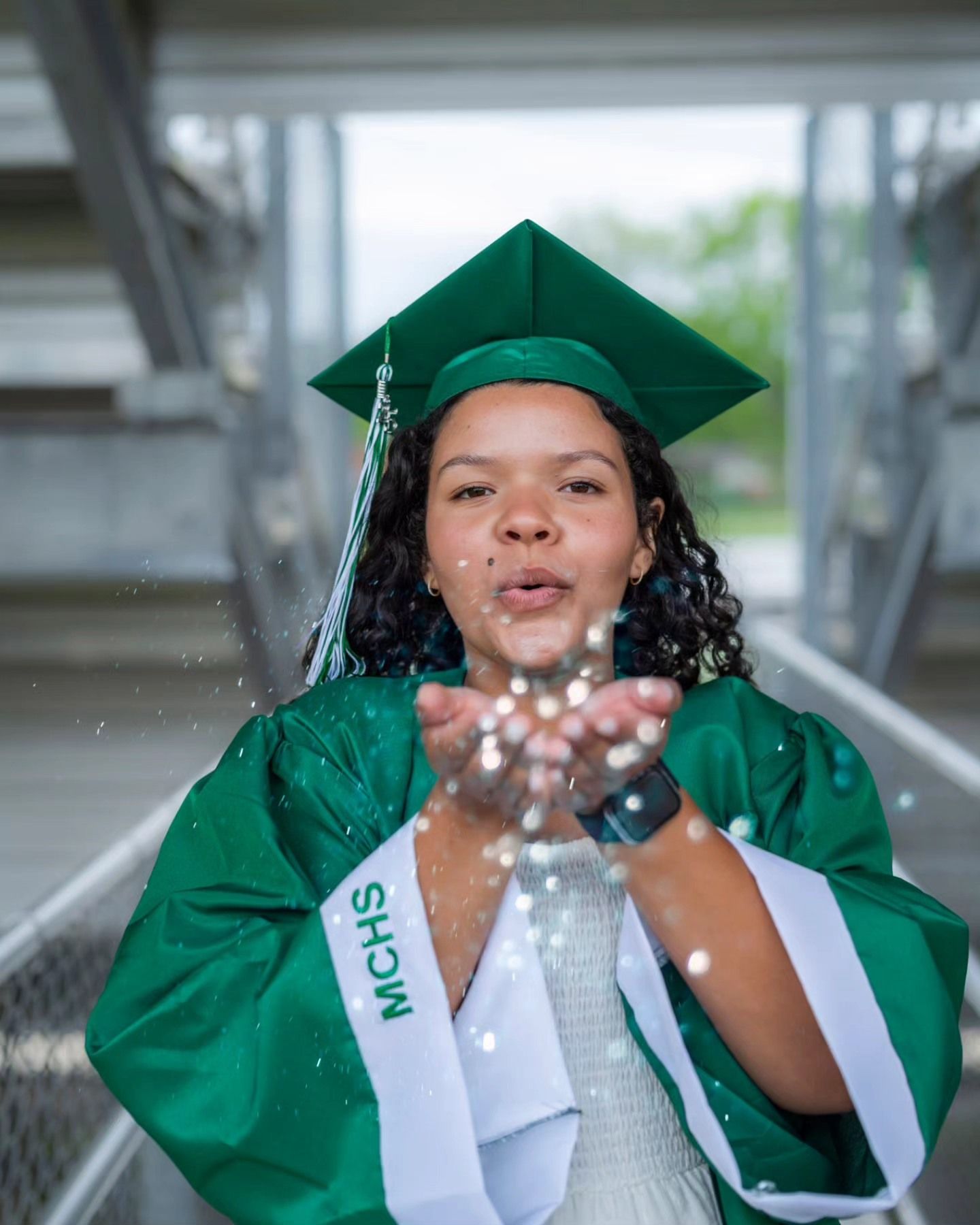 Female graduate in green MCHS cap and gown blows glitter toward the camera.