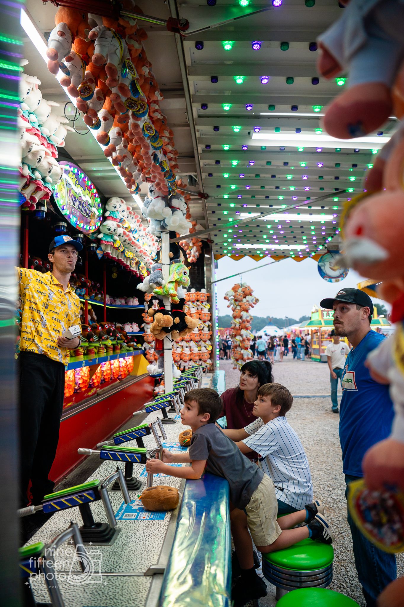 Kids play a carnival game at a colorful fair midway booth with stuffed animal prizes and festive lights.