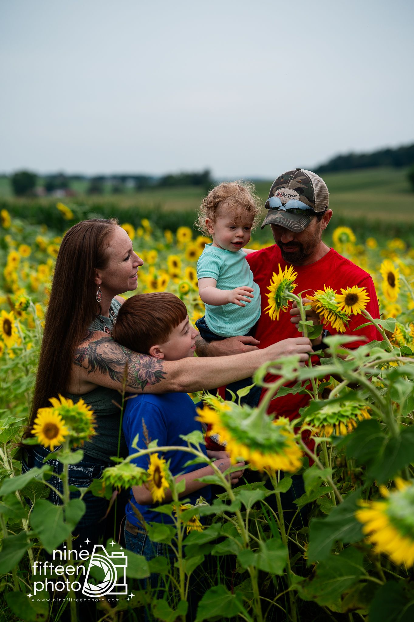 Family of four enjoying a sunflower field, with parents holding young children while admiring blooming sunflowers on a cloudy day.