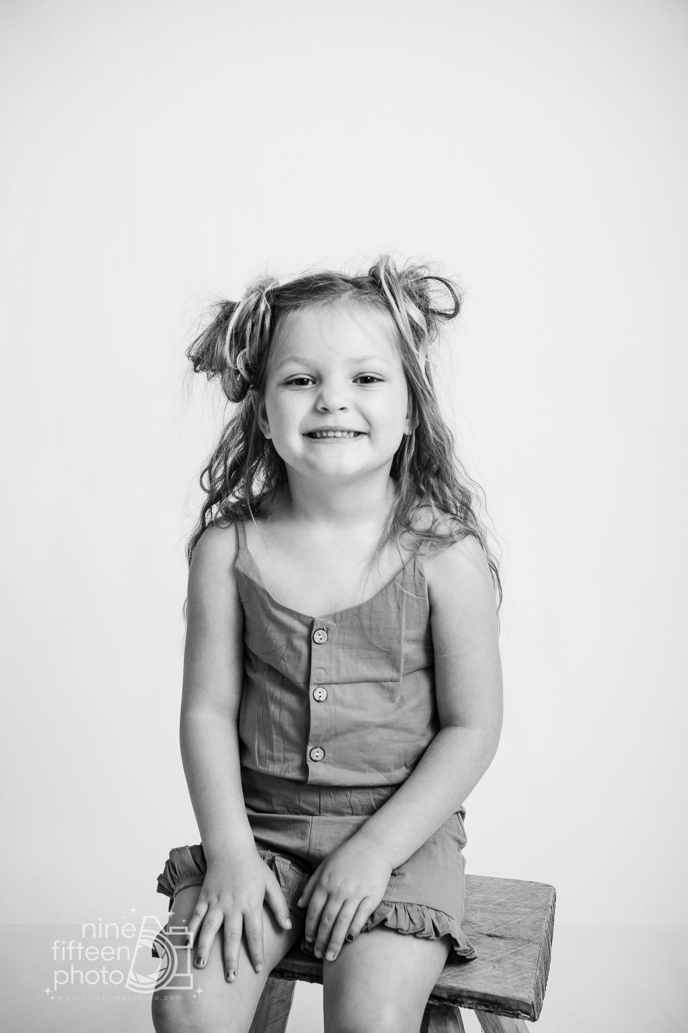 Black and white portrait of a smiling young girl with pigtails sitting on a wooden stool wearing a romper.