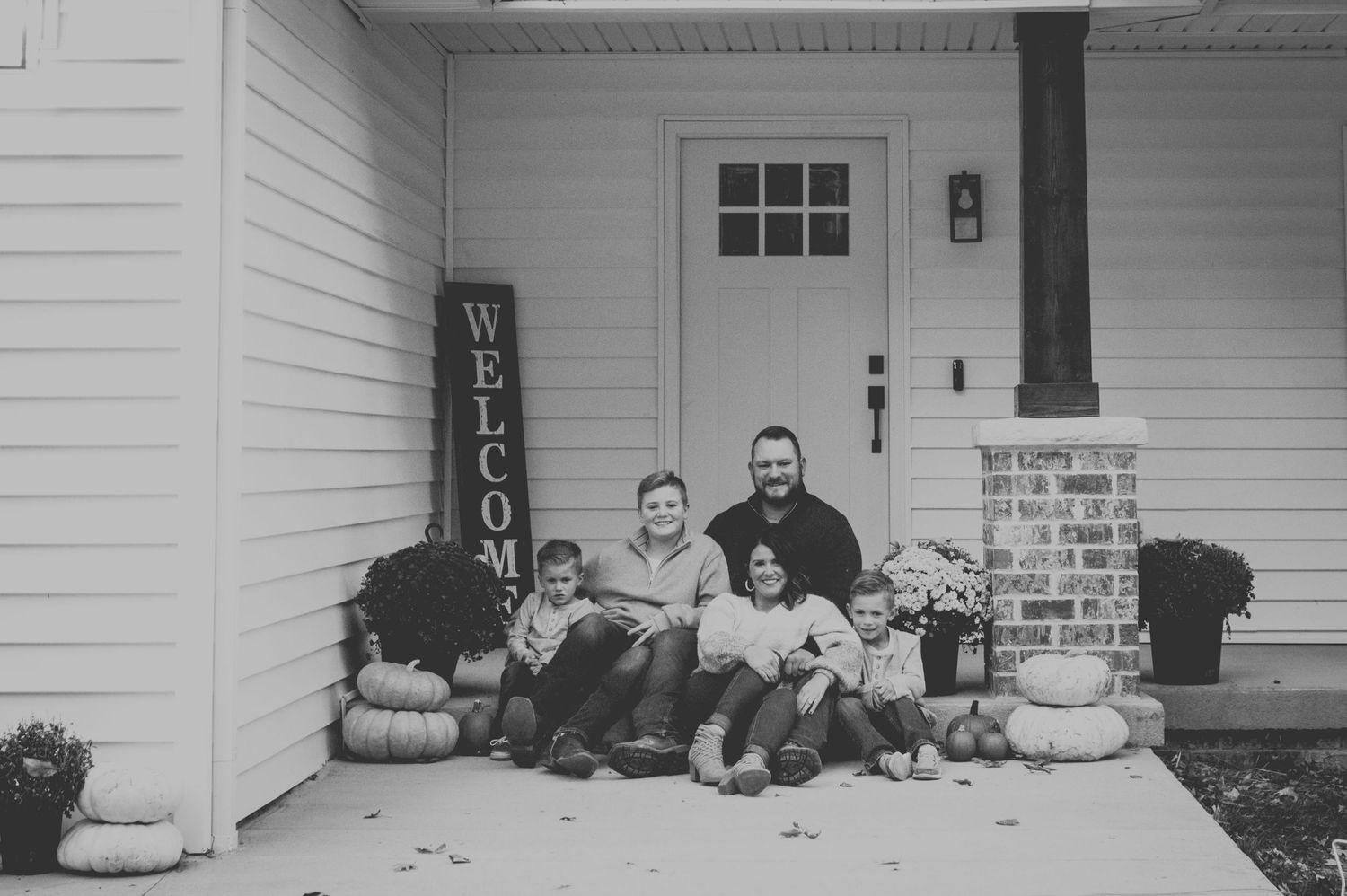Black and white photo of a family sitting on front porch steps with pumpkins and a welcome sign in fall.