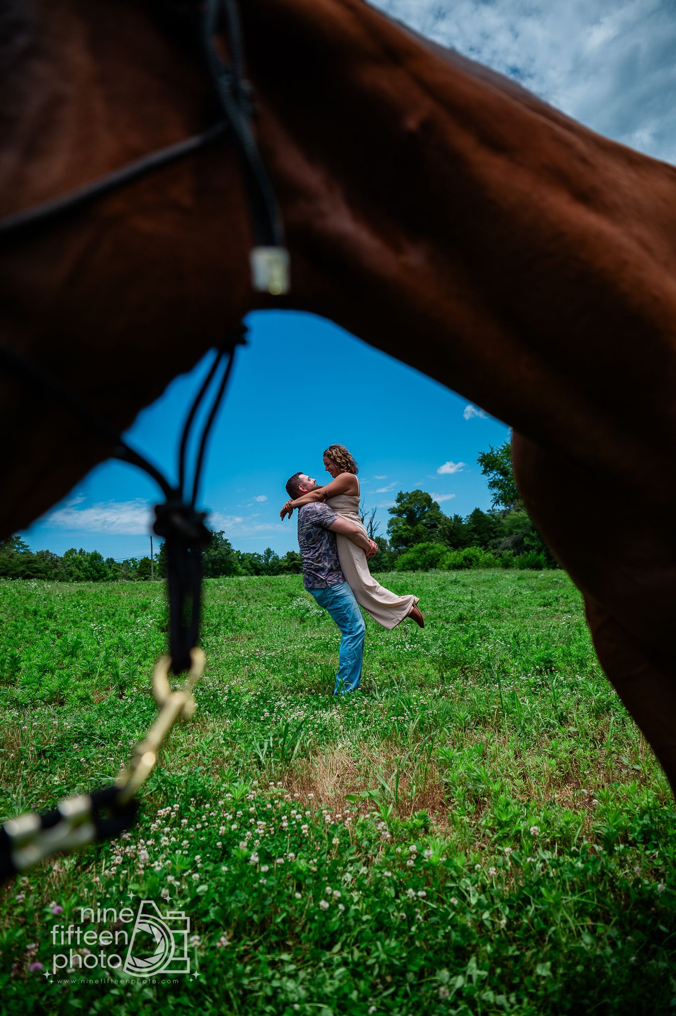 Couple embracing in green field photographed through horse legs under blue sky, romantic engagement photo by Nine Fifteen Photo.