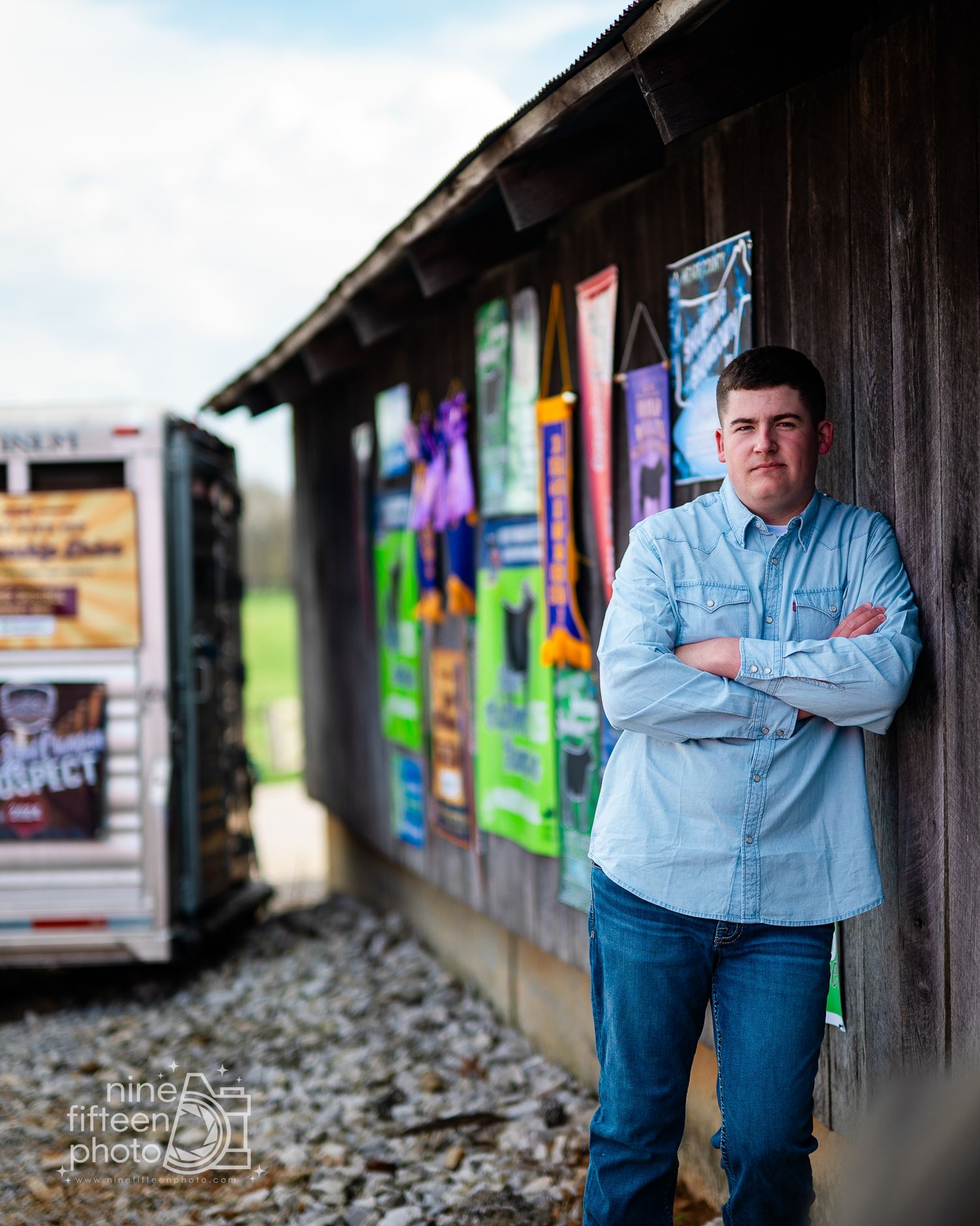 Young man in light blue shirt stands arms crossed against rustic wooden wall with colorful posters outdoors.