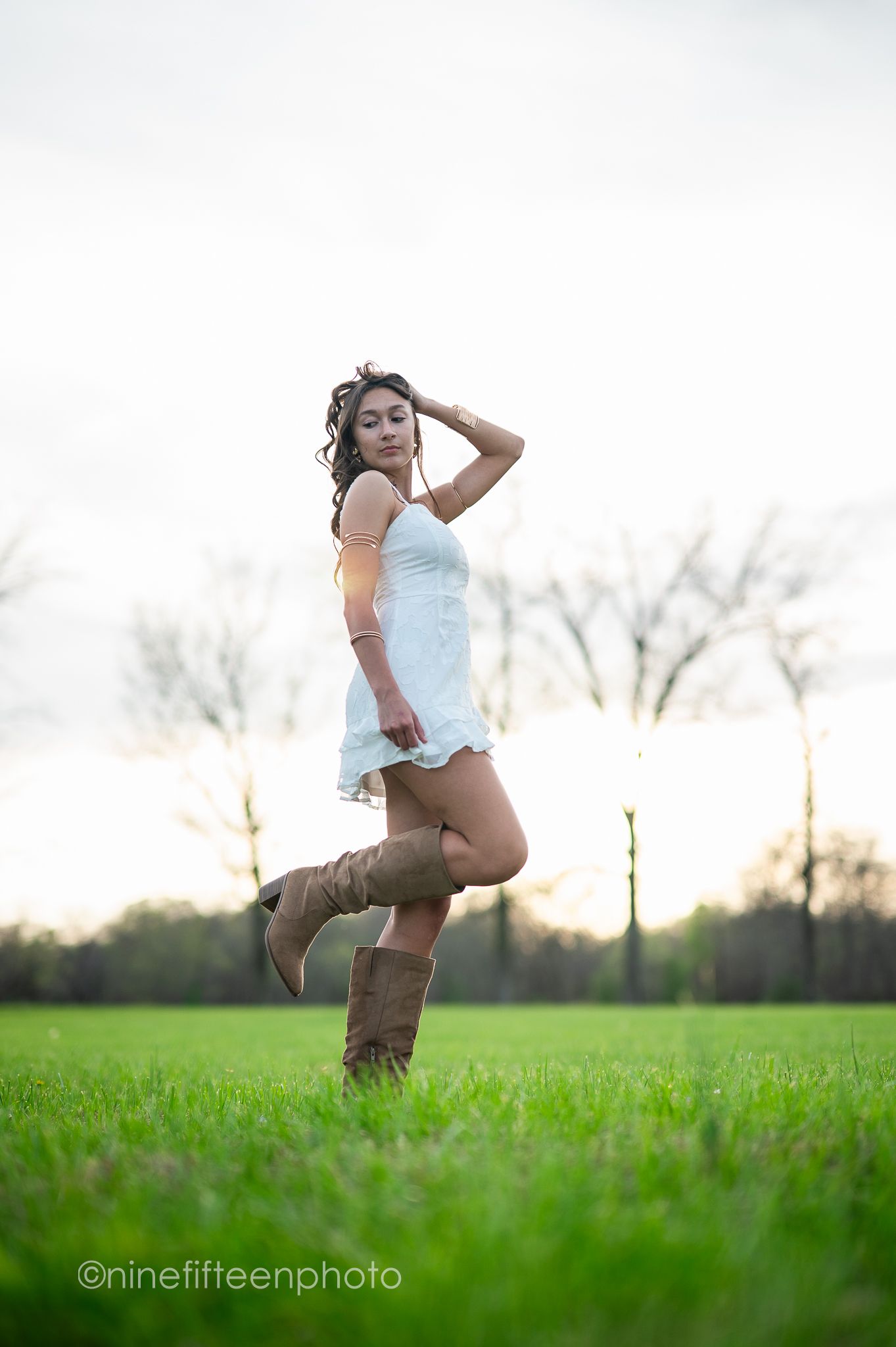 Young woman in a white dress and brown cowboy boots posing playfully in a green field at dusk.