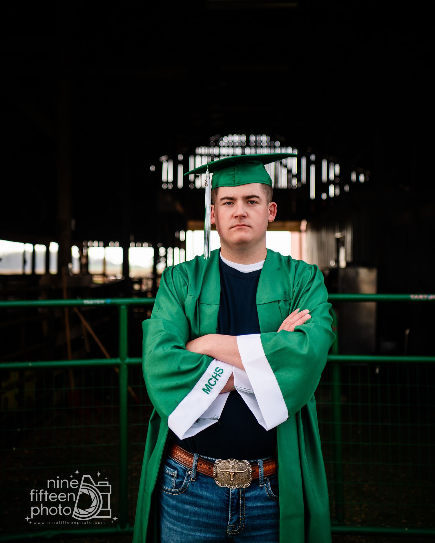 Young man in green graduation cap and gown with white stole poses confidently with arms crossed outdoors.