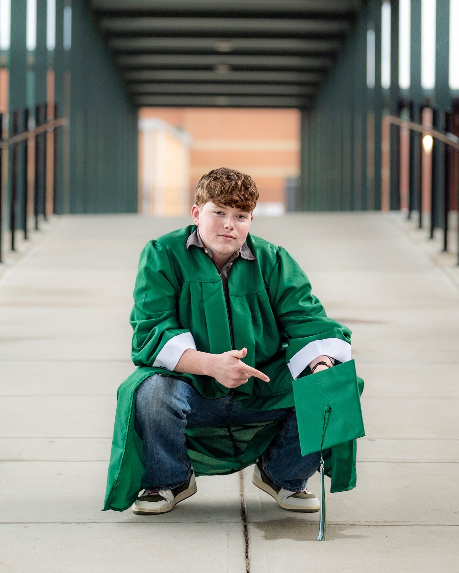 Male graduate in green gown crouches on a bridge holding his cap in a senior portrait.