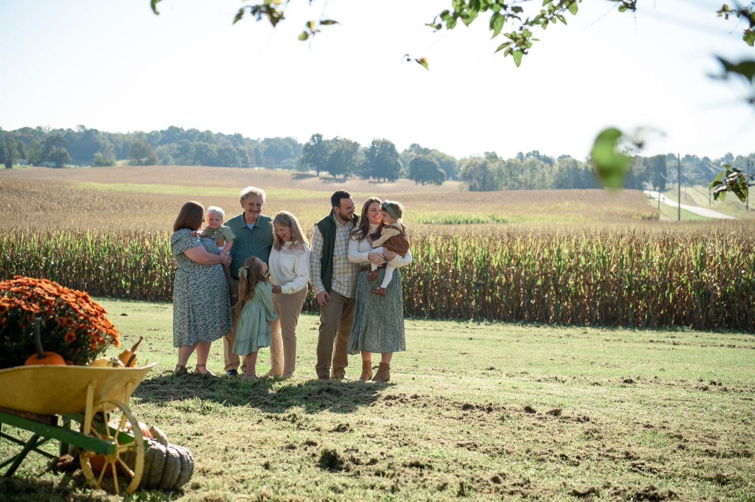 Extended family group portrait outdoors near a cornfield with orange mums and a wooden wagon on a sunny fall day.