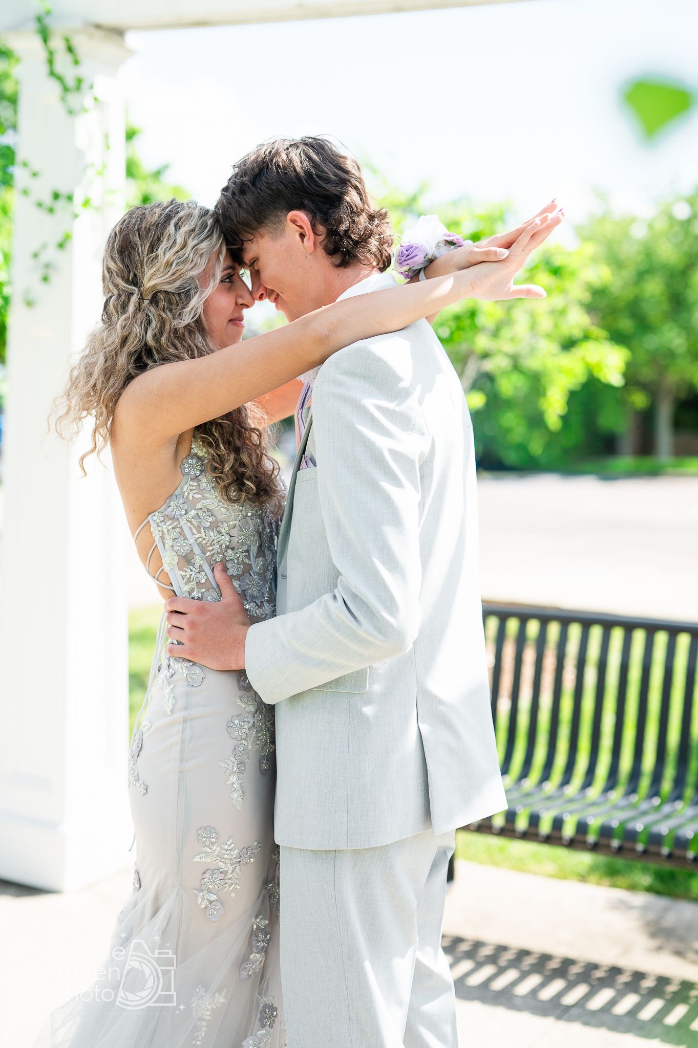 Couple in formal attire shares a romantic embrace under a white gazebo during prom or wedding.