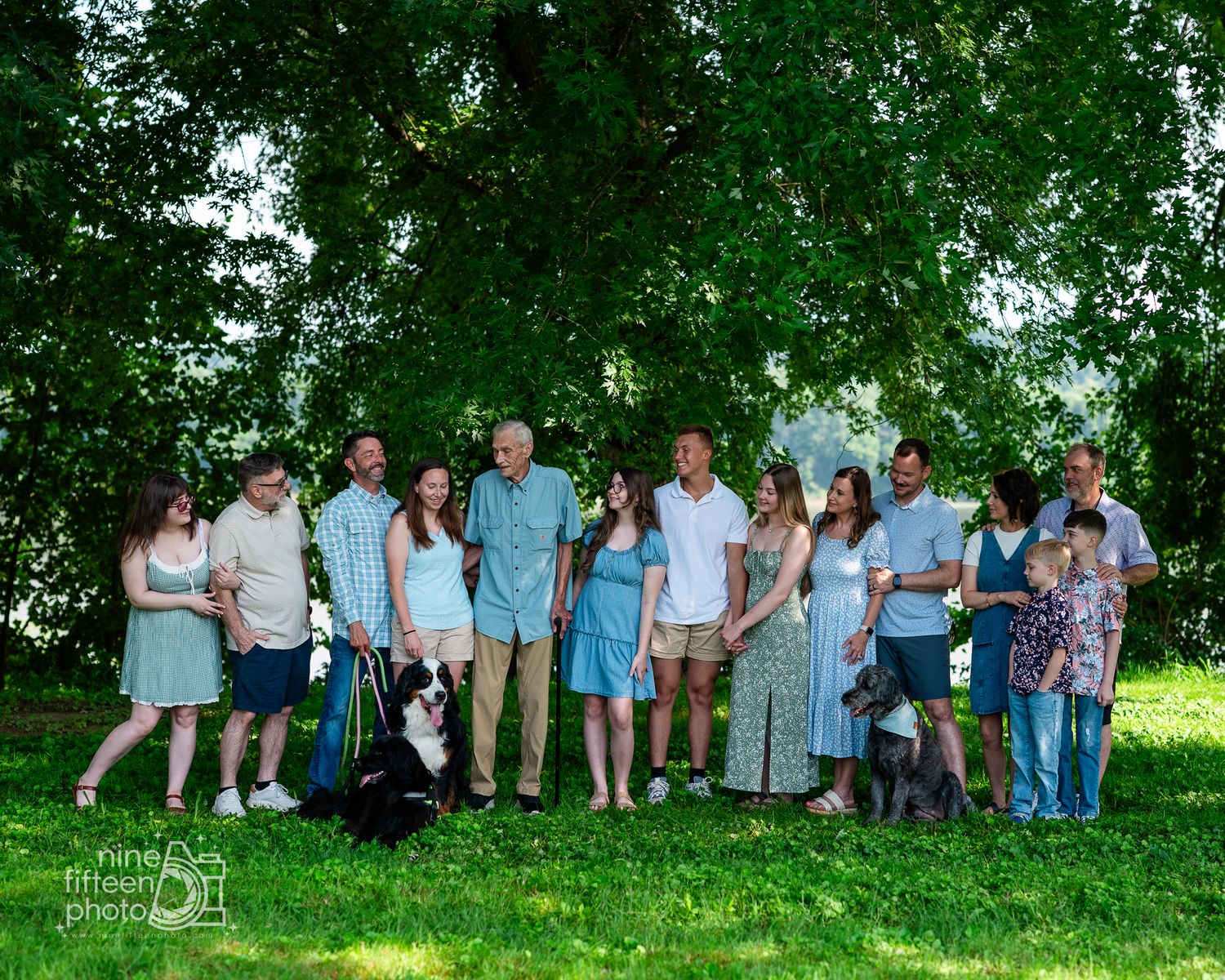 Large family group portrait outdoors under lush green trees on a sunny day, including adults, children, and a dog.