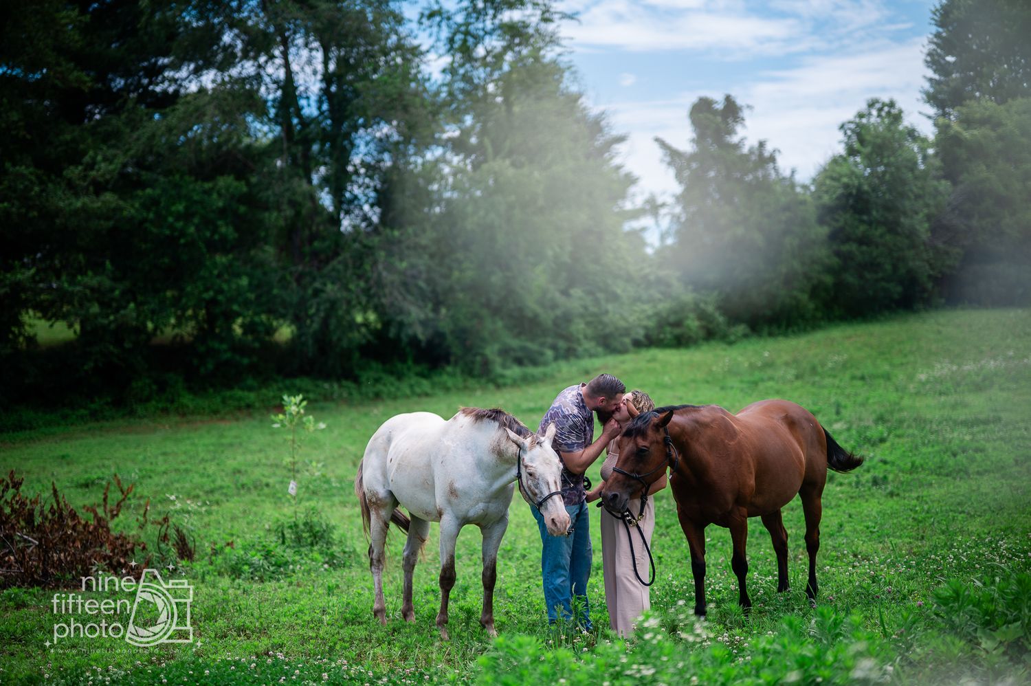 Person standing in green field with two horses, one white and one brown, surrounded by lush trees on a misty day.