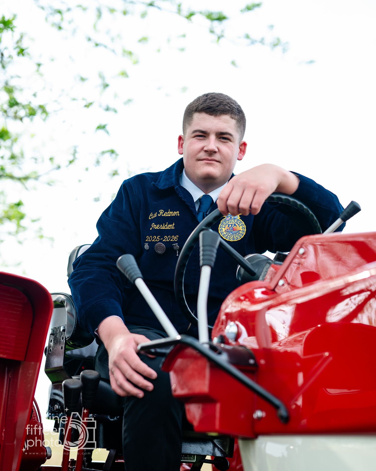 Young man in FFA blue jacket sitting on a red tractor, wearing a president badge, posed for a senior portrait outdoors.