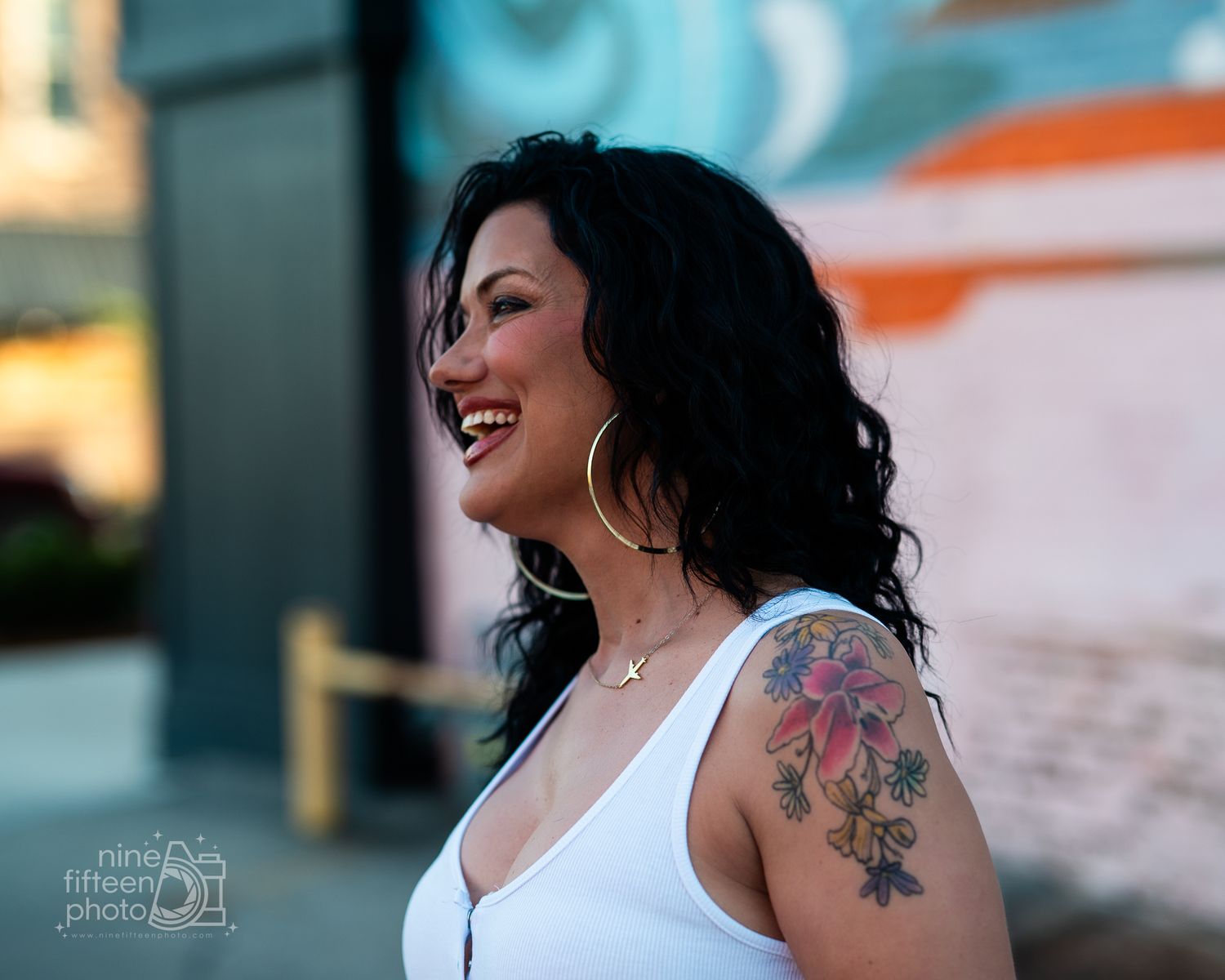 Laughing woman with black hair, hoop earrings, and floral shoulder tattoo poses outdoors in white top.