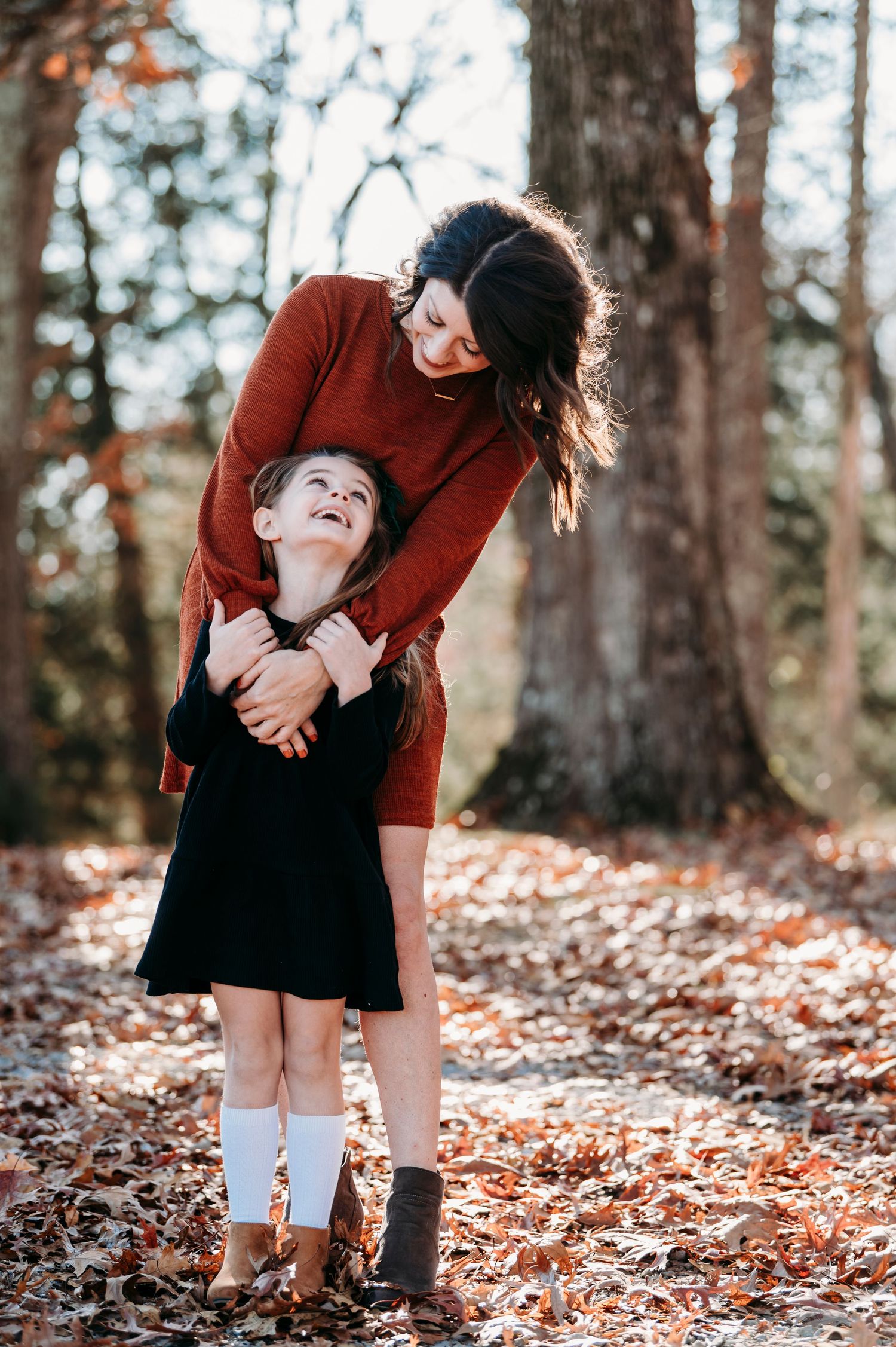 A woman in a rust dress hugs a laughing girl in a black dress among autumn leaves in a forest.