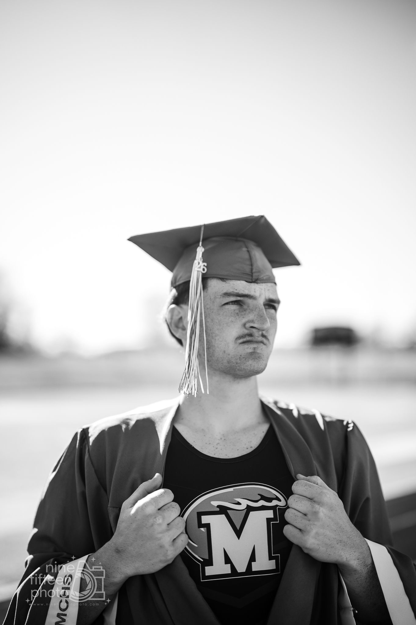 Black and white senior portrait of male graduate in cap and gown revealing varsity M shirt.