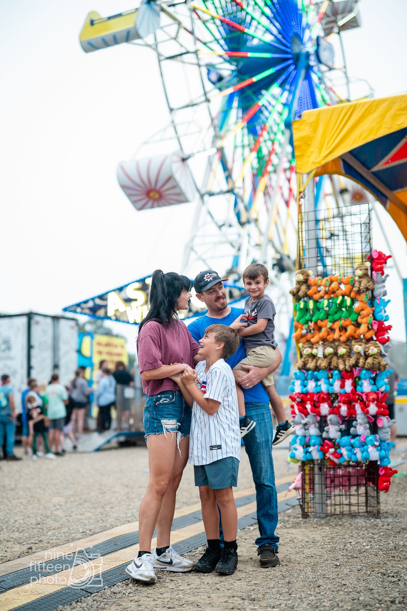 Family of four smiling together at a colorful county fair with a Ferris wheel and prize stuffed animals in the background.