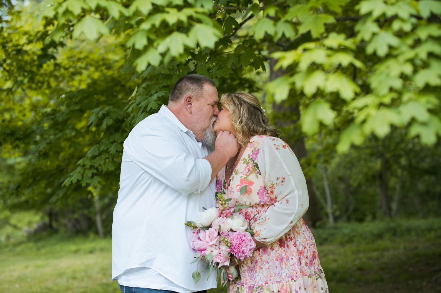 Couple sharing a romantic kiss outdoors under lush green trees, woman holding pink bouquet in floral dress.