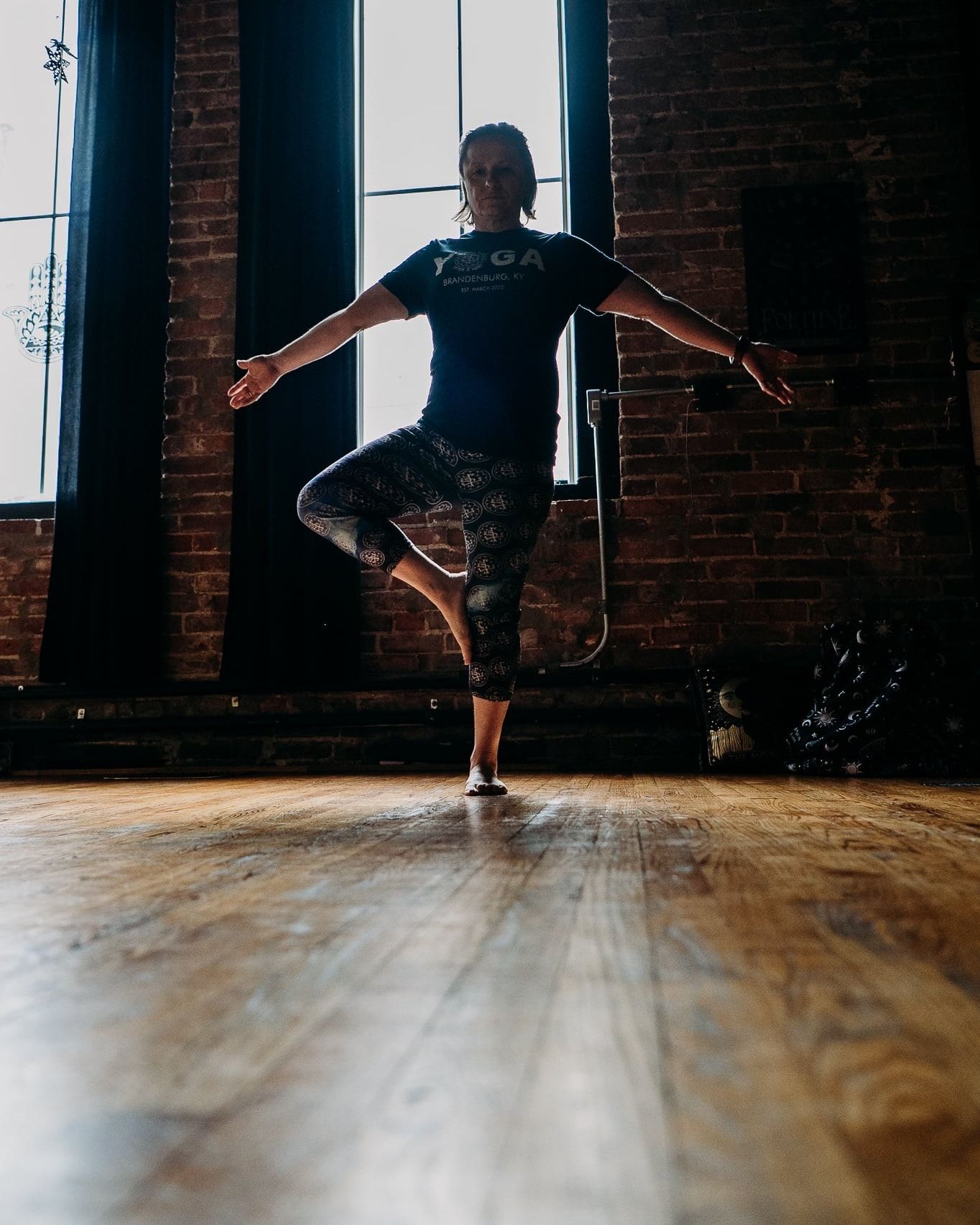Woman practicing tree pose yoga on hardwood floor in front of brick wall and large window.
