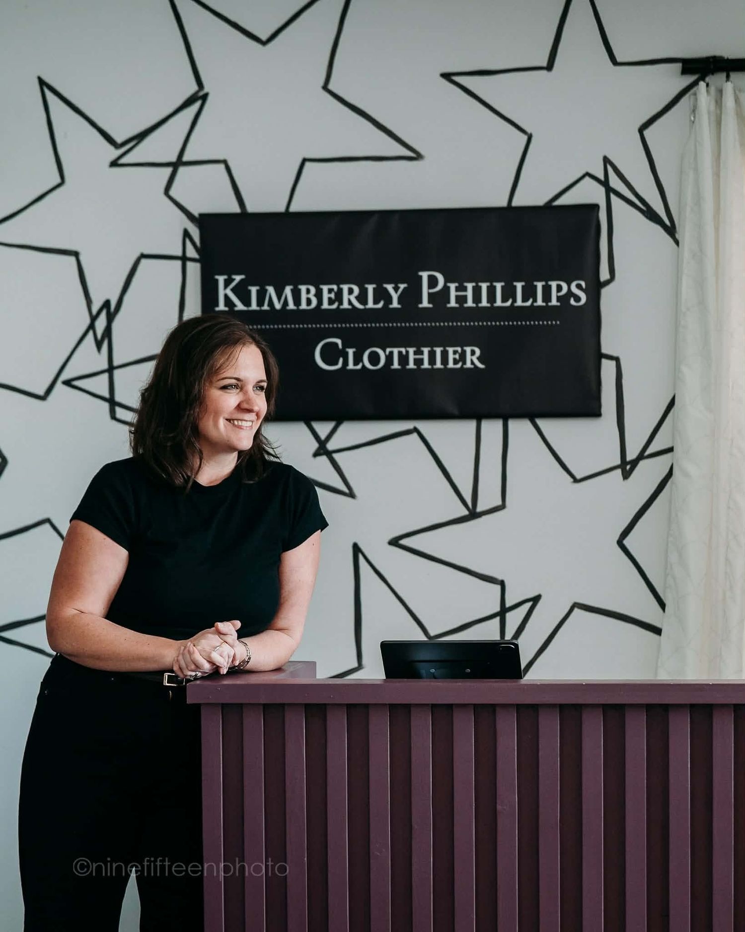Kimberly Phillips Clothier owner smiling behind purple counter in bridal boutique with star decor.