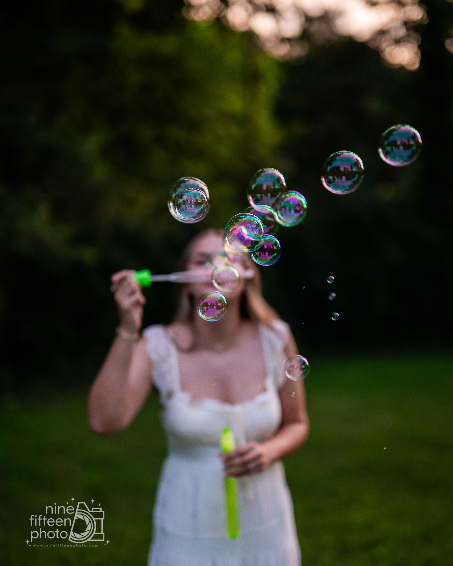 A young woman in a white dress blows colorful soap bubbles outdoors in a lush green park at dusk.