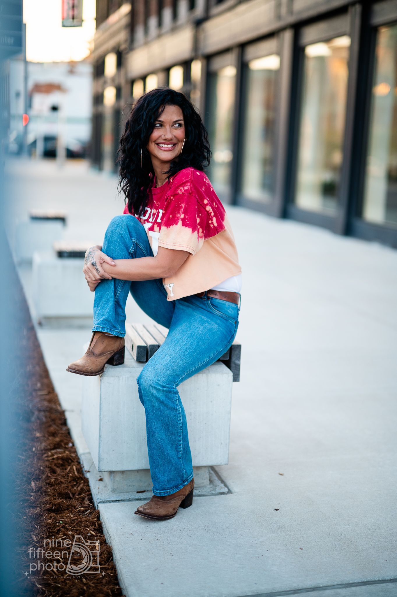 Woman in pink tie-dye shirt and jeans sitting on concrete bench outdoors, smiling, wearing cowboy boots.