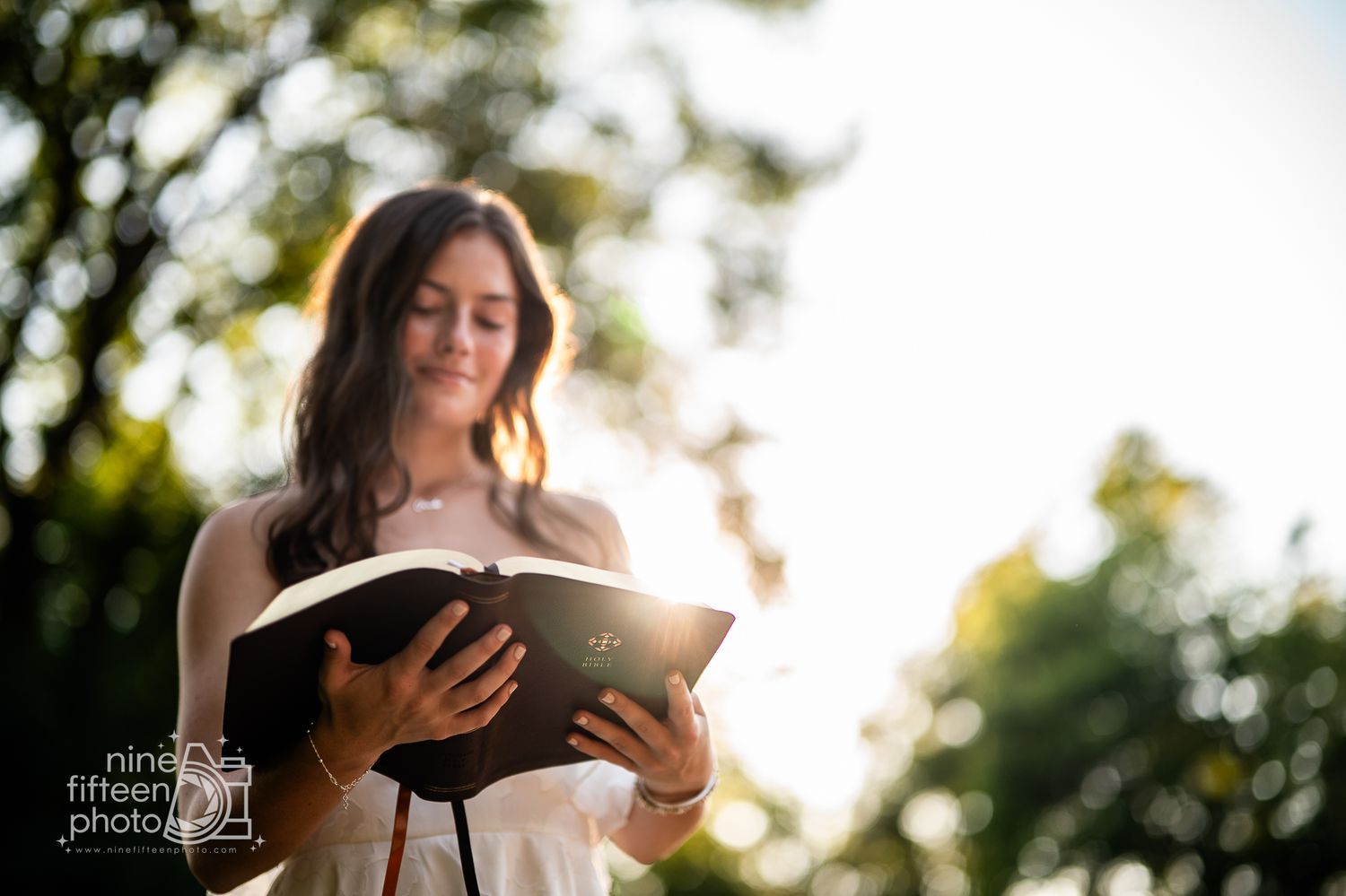 Young woman with long brown hair reads a book outdoors with sunlight filtering through trees behind her.