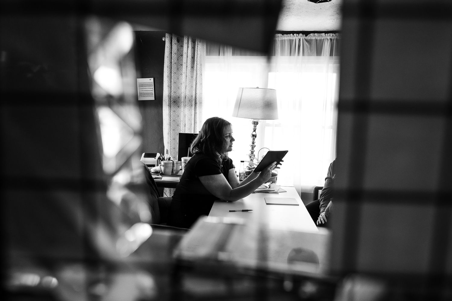 Woman reading a tablet at a desk by a window, captured through blinds in black and white.
