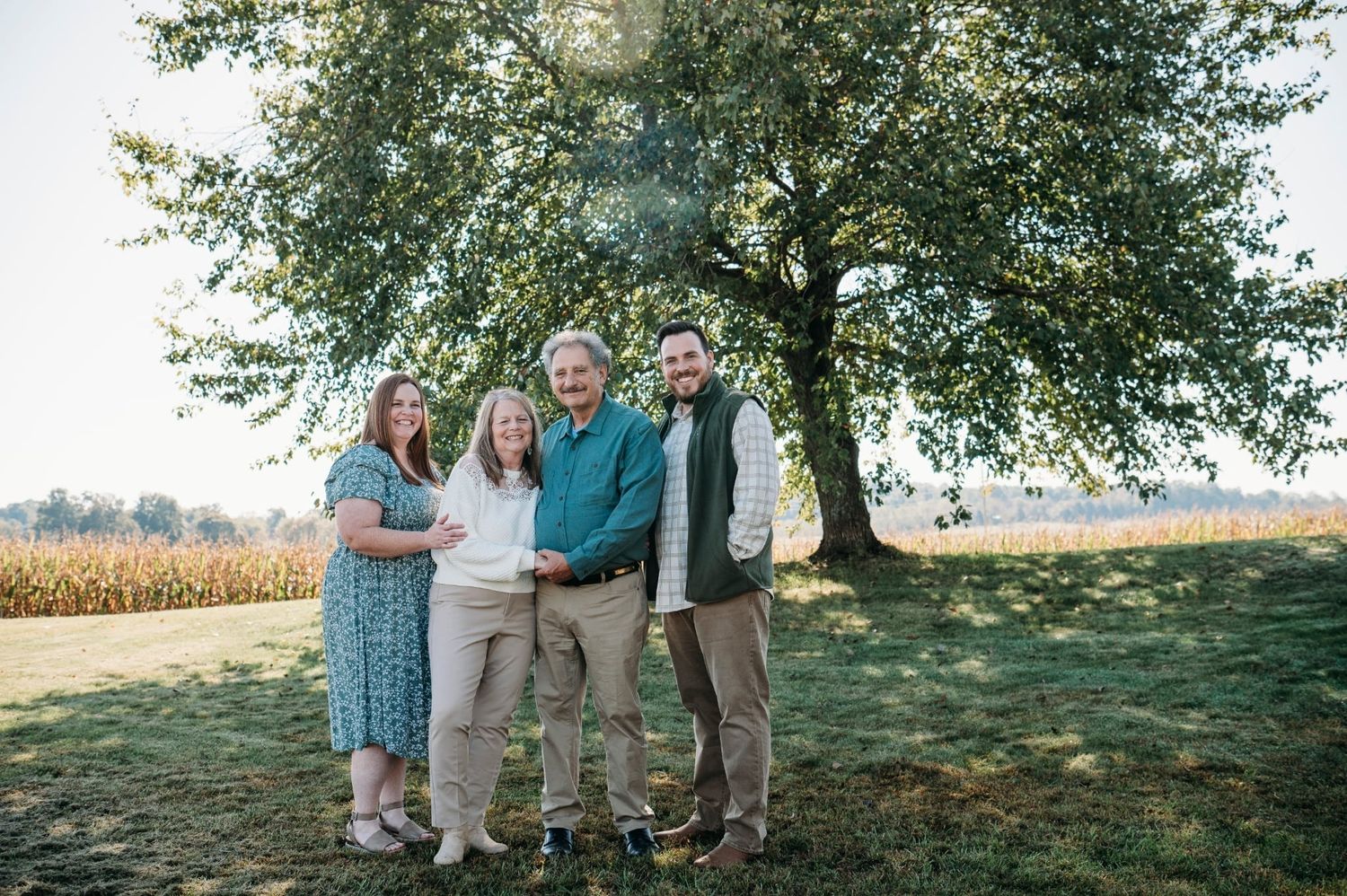 Family of four smiling together outdoors near a large tree with a cornfield in the background on a sunny day.